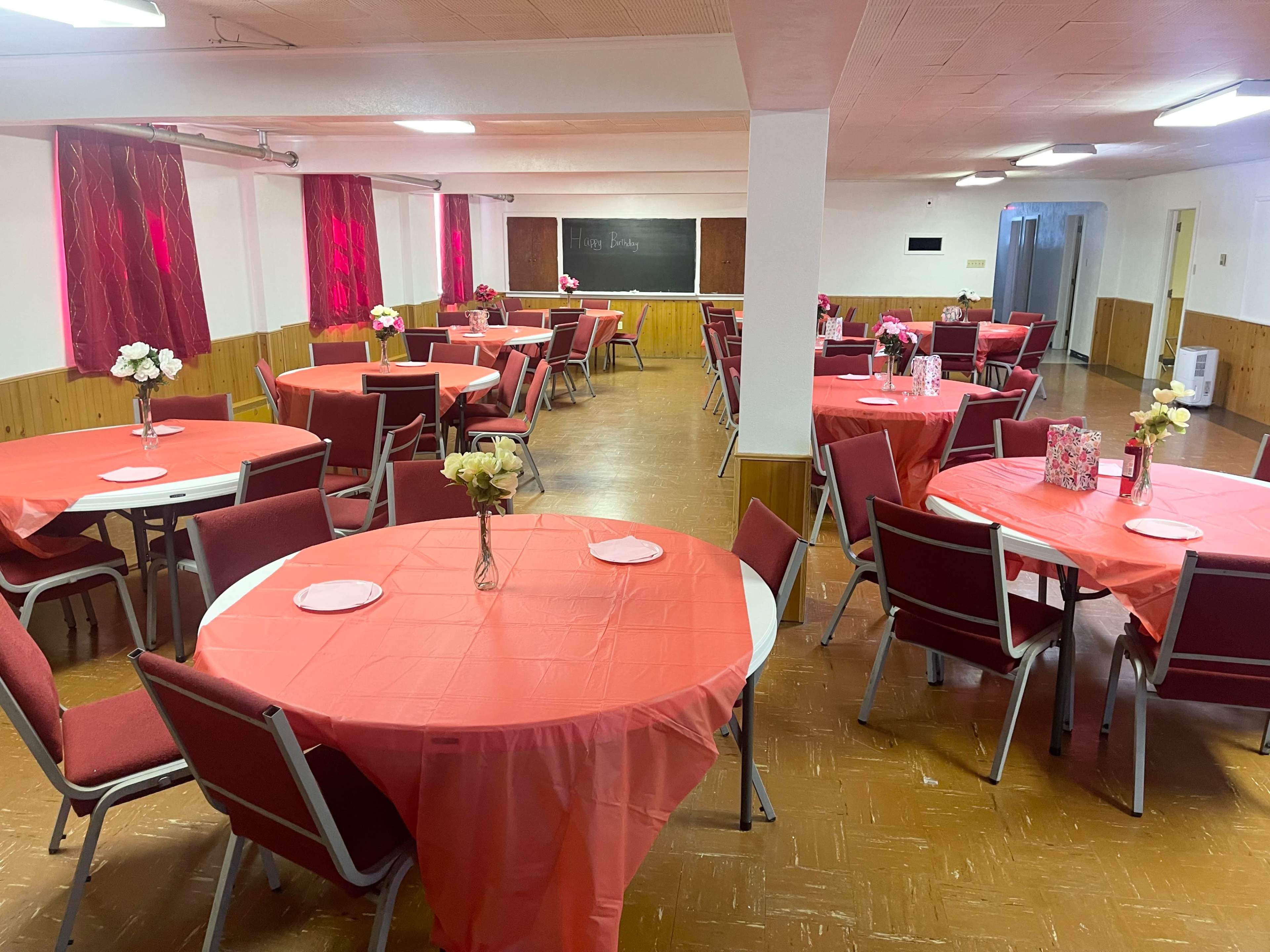A spacious room set up for an event features round tables covered with red tablecloths and decorated with flowers, with additional seating arranged against the walls.