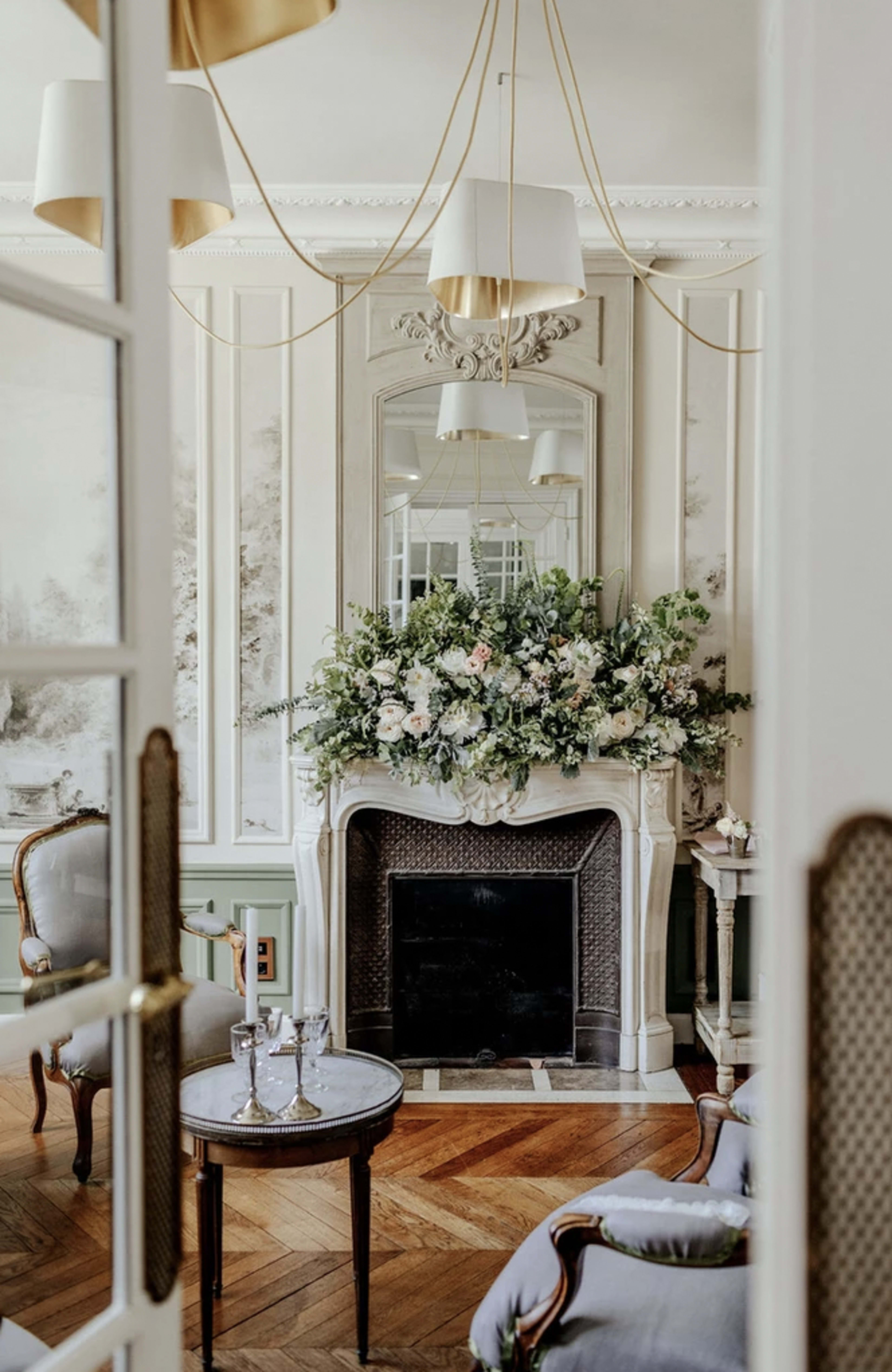 A well-decorated living room features a fireplace adorned with a floral arrangement, elegant furnishings, and a stylish chandelier.