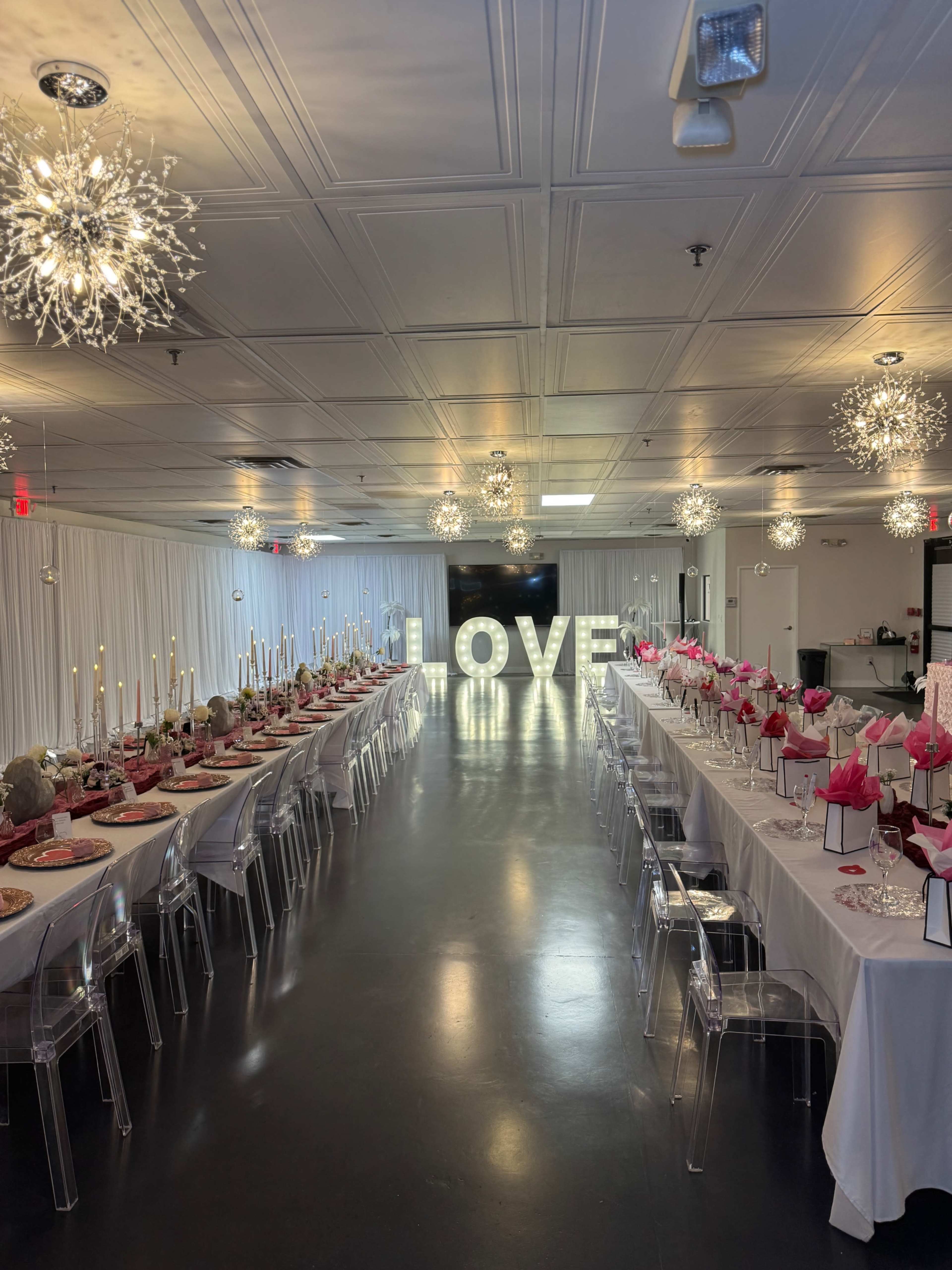 The image shows a banquet hall arranged with two long tables decorated with pink accents and a large illuminated "LOVE" sign in the center.