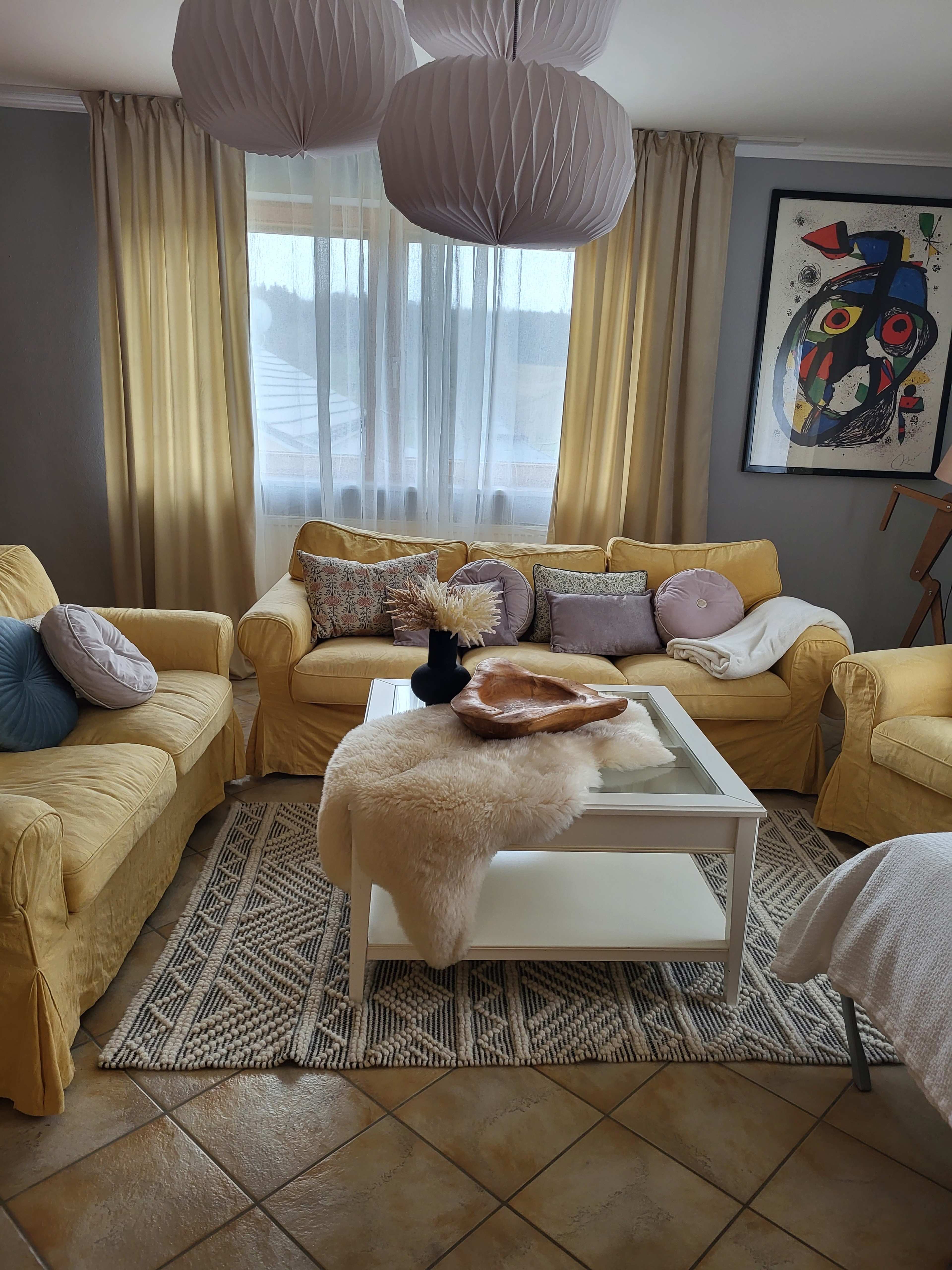 A bright living room features yellow sofas, a glass coffee table with decorative items, and large light-filtering curtains.