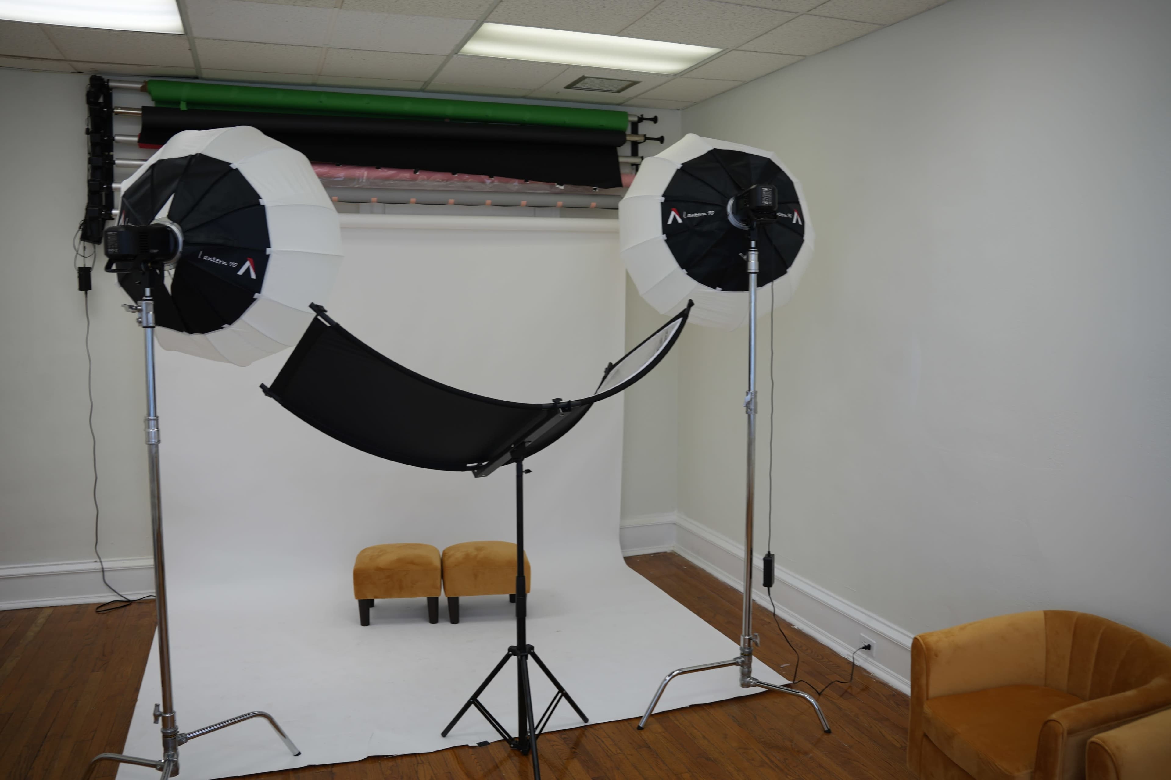 A photography studio with two softbox lights, a black backdrop support, and two small stools positioned in front of a white backdrop.