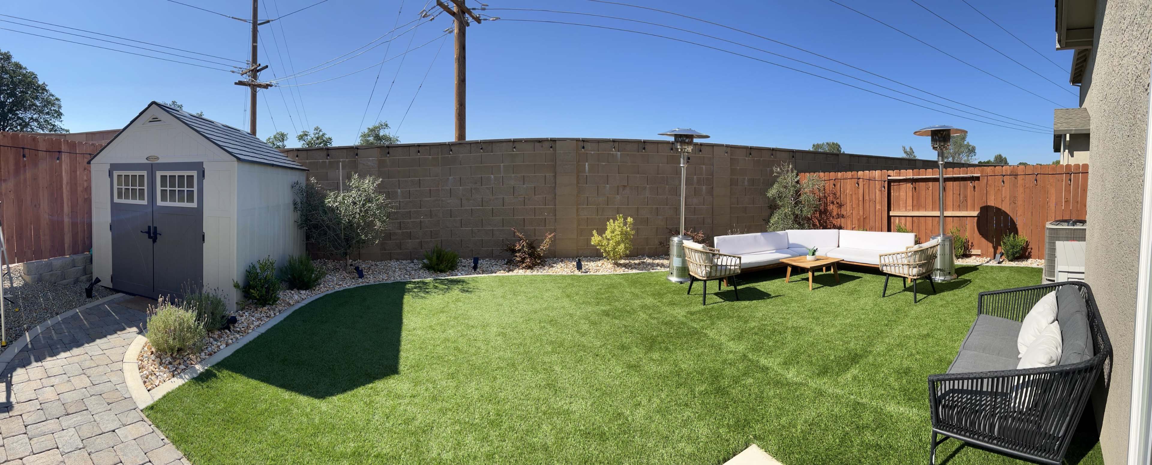 A landscaped backyard featuring artificial grass, a seating area with sofas and chairs, a small shed, and outdoor heaters surrounded by a wooden fence.