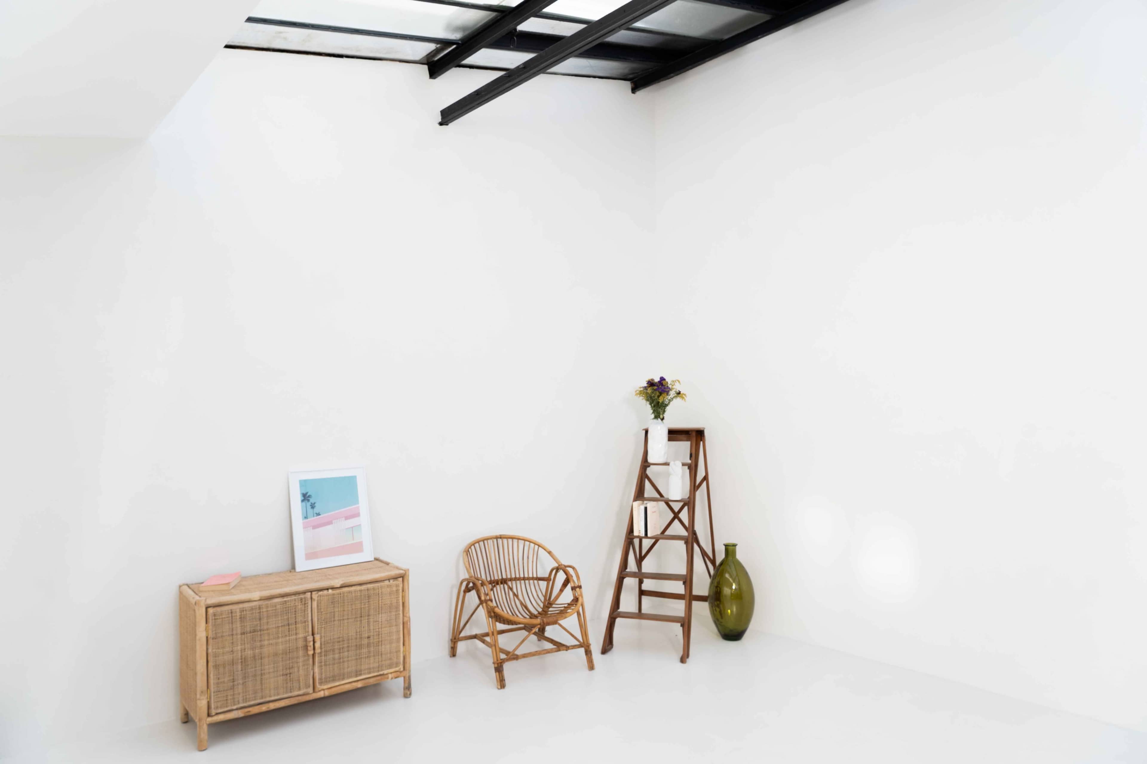 The image shows a minimalist interior space featuring a wicker chair, a wooden side cabinet, a ladder-style shelf with a vase, and a framed picture against a white wall.