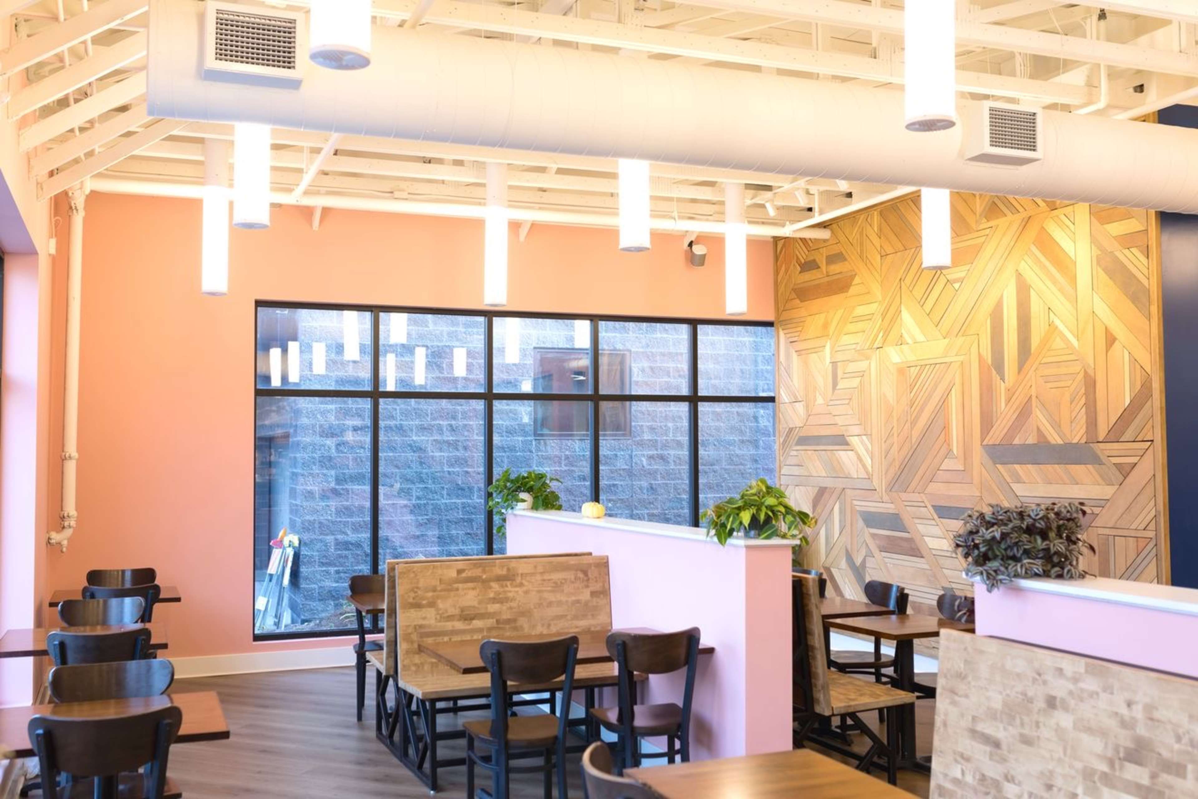 The image shows the interior of a cafe featuring wooden tables, pink walls, and large windows allowing natural light to enter.