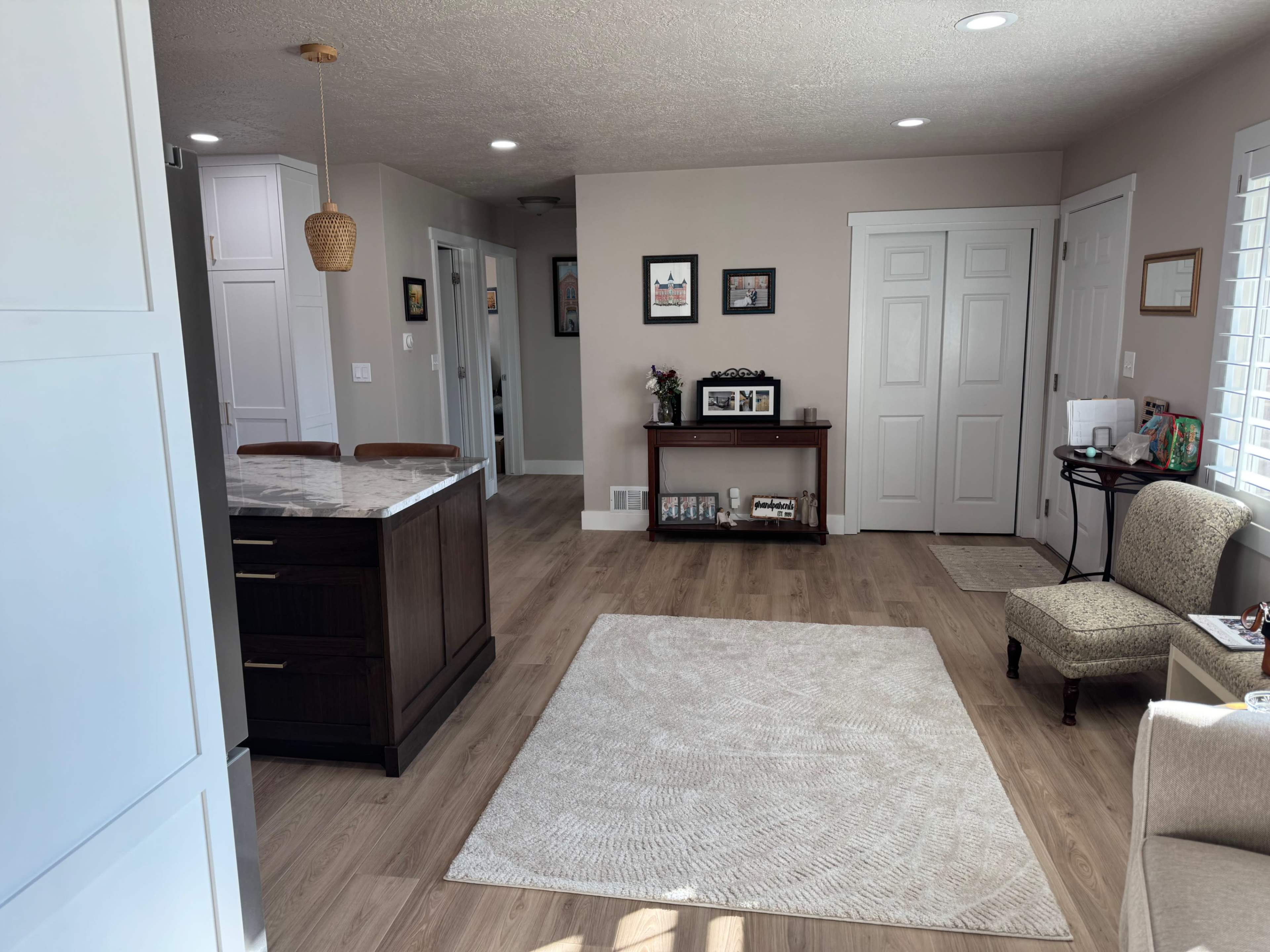 The image shows a well-lit living room and kitchen area with a central rug, a dining island with stools, and a sideboard against the wall.