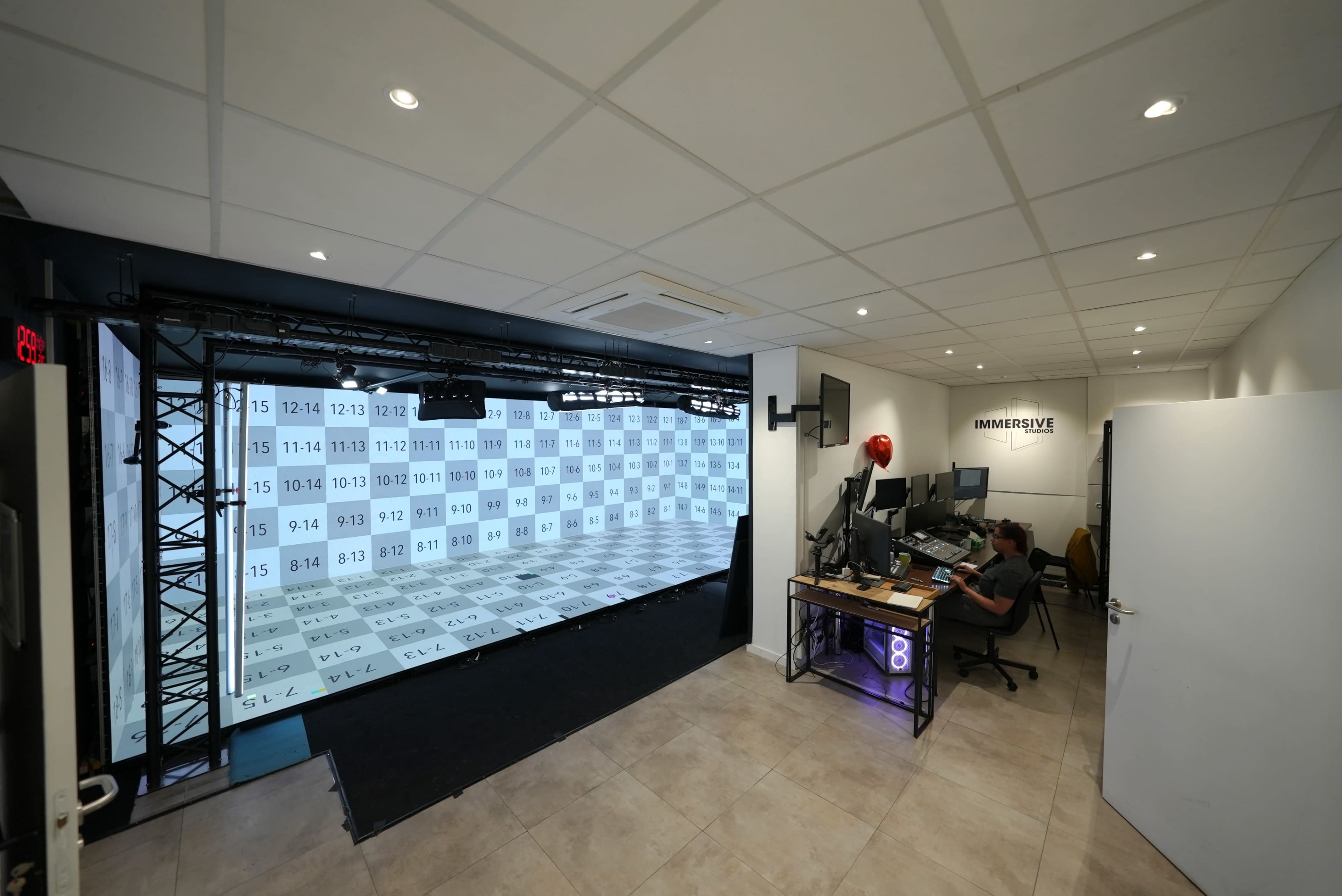 The image shows a modern office space featuring a large projection area with grid patterns on the wall and a desk with multiple computer screens in a well-lit environment.