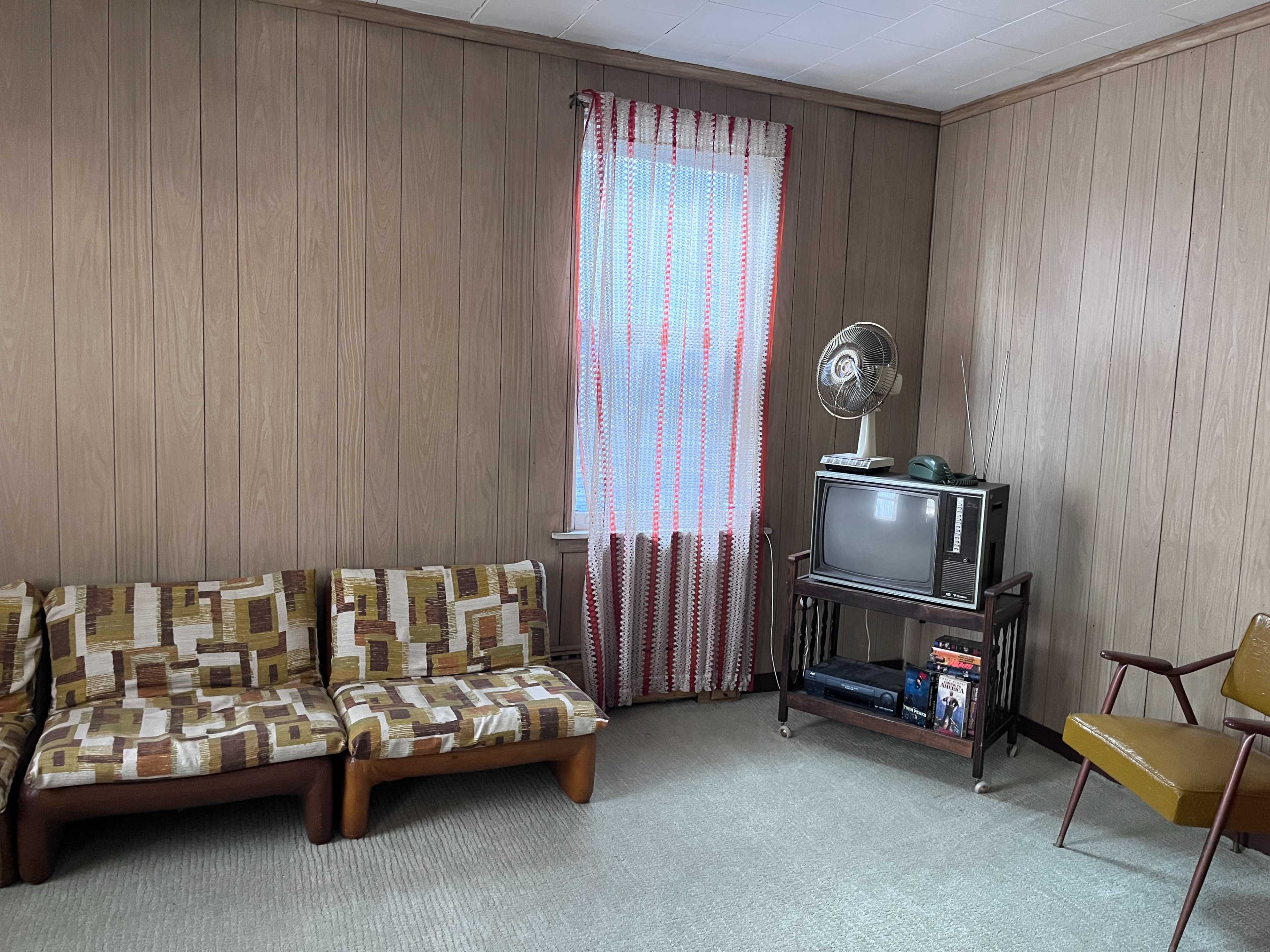 The image shows a small, sparsely furnished room with wood-paneled walls, a patterned couch, a standing fan, a vintage television on a cart, and a chair.