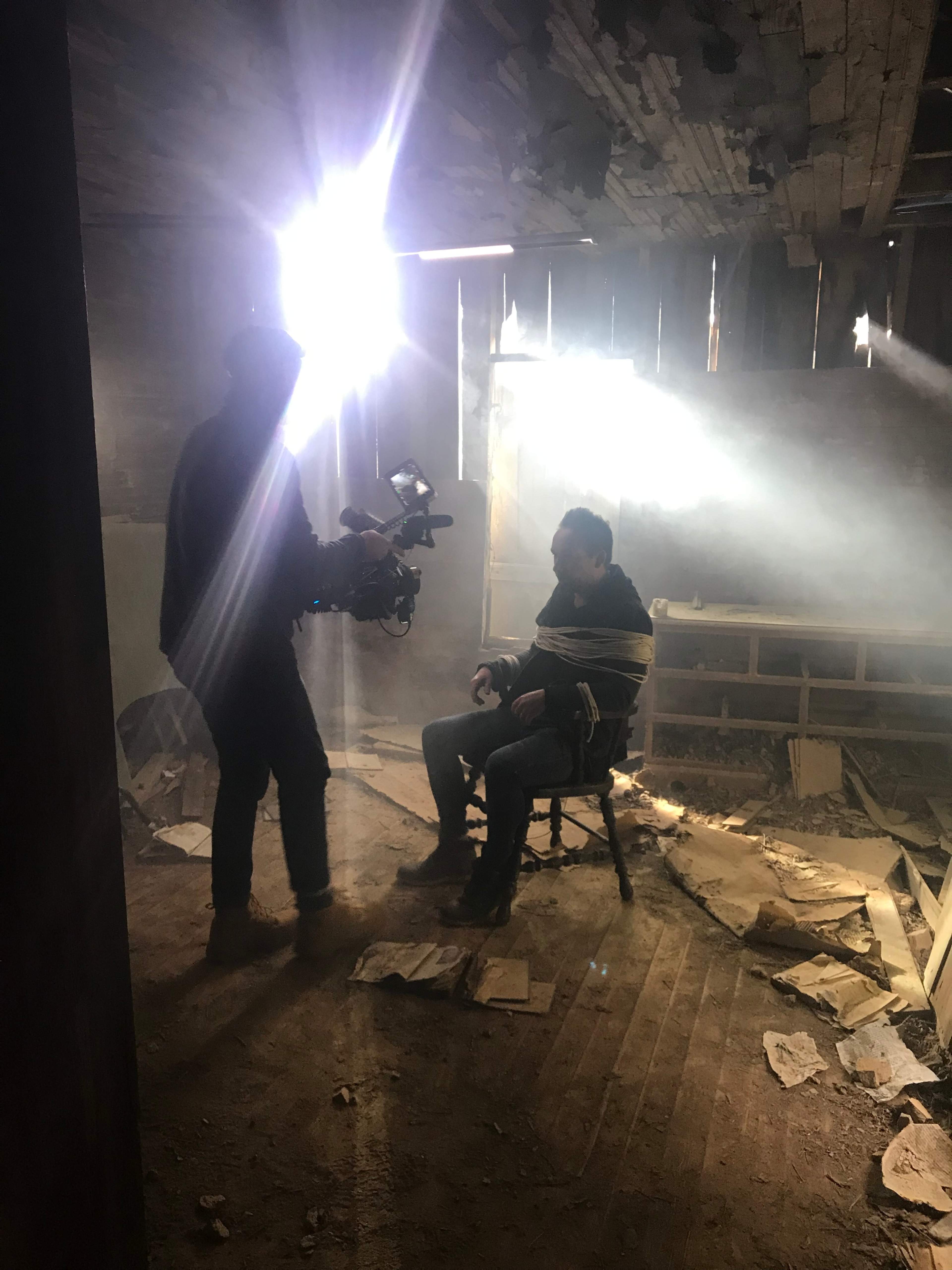 A man is tied to a chair in a dimly lit room while a person uses a camera to film him.