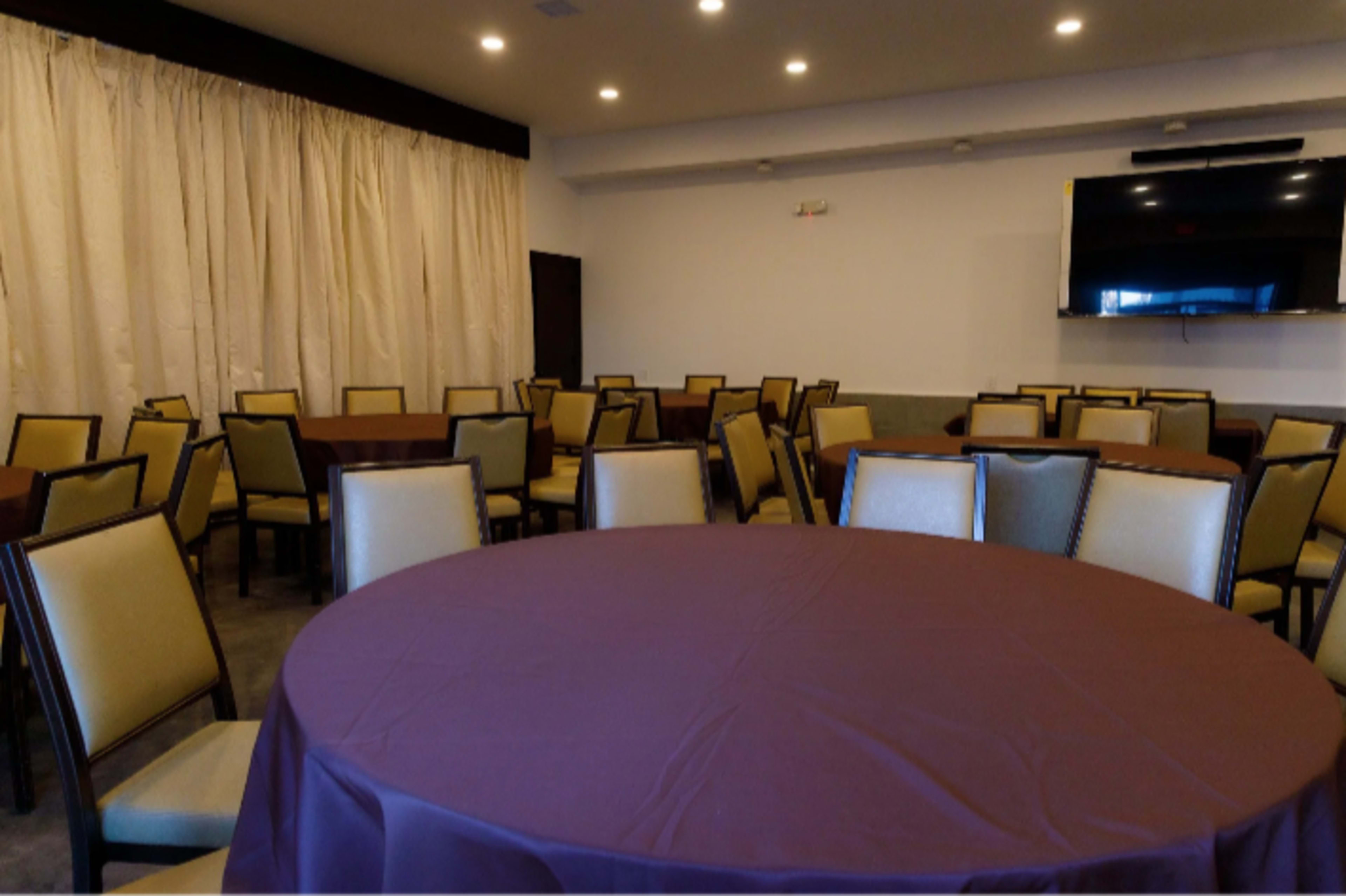The image shows a spacious indoor room with multiple sets of round tables, each covered with brown tablecloths, surrounded by yellow chairs and a large screen on the wall.