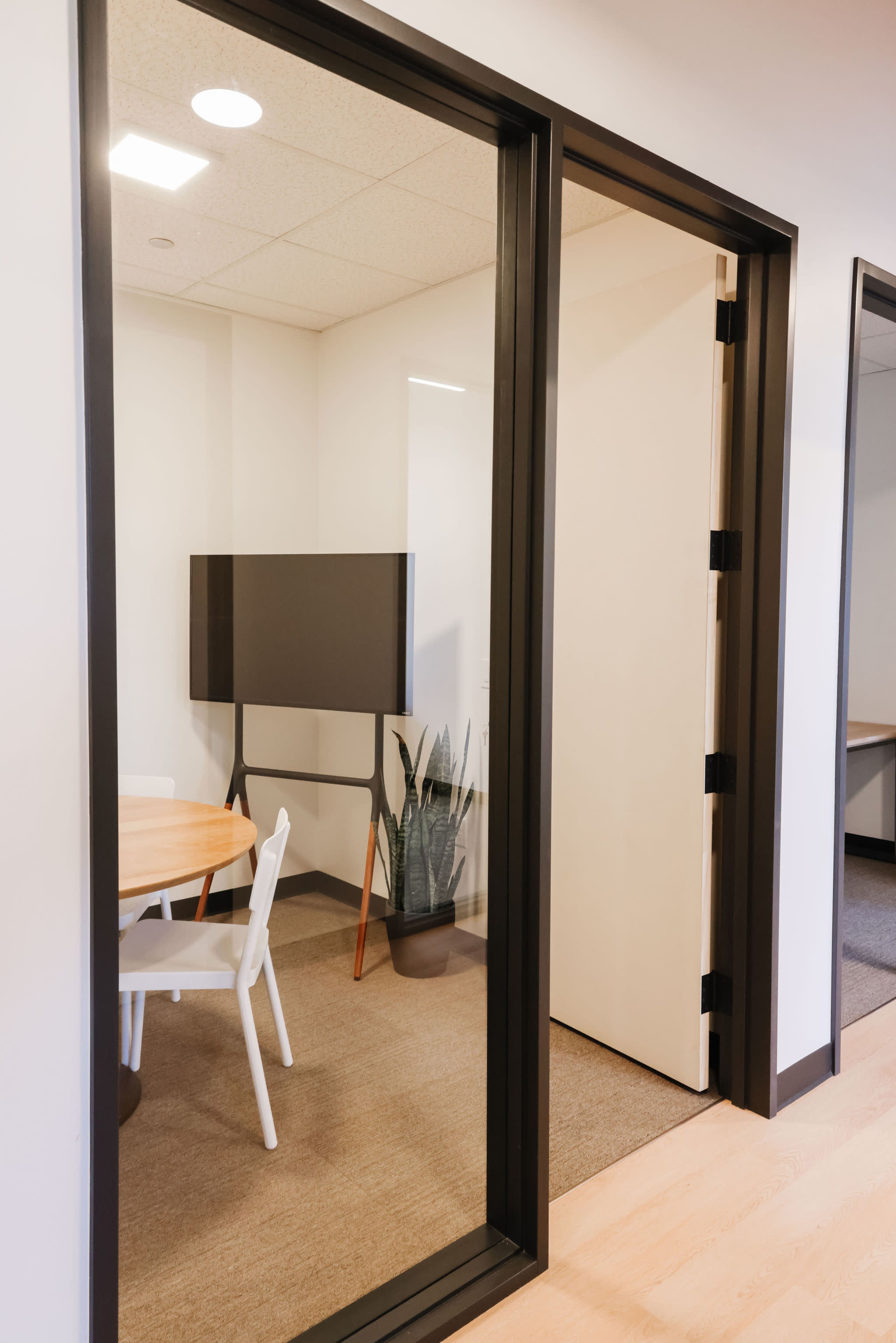 A modern office space featuring a glass-walled meeting room with a round table, white chairs, and a plant.