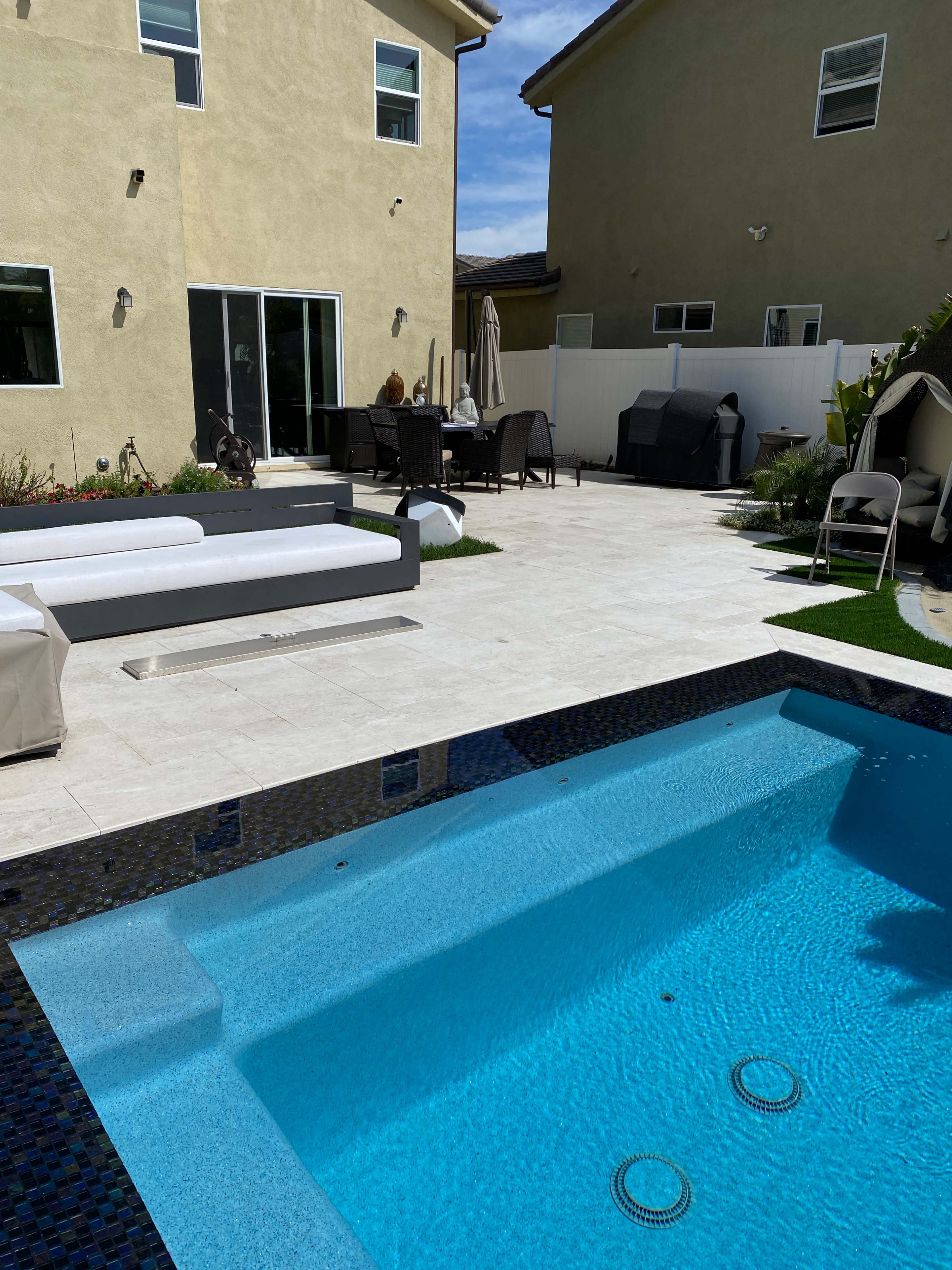 A backyard patio with a swimming pool, modern outdoor furniture, and a dining area adjacent to a green lawn and fence.