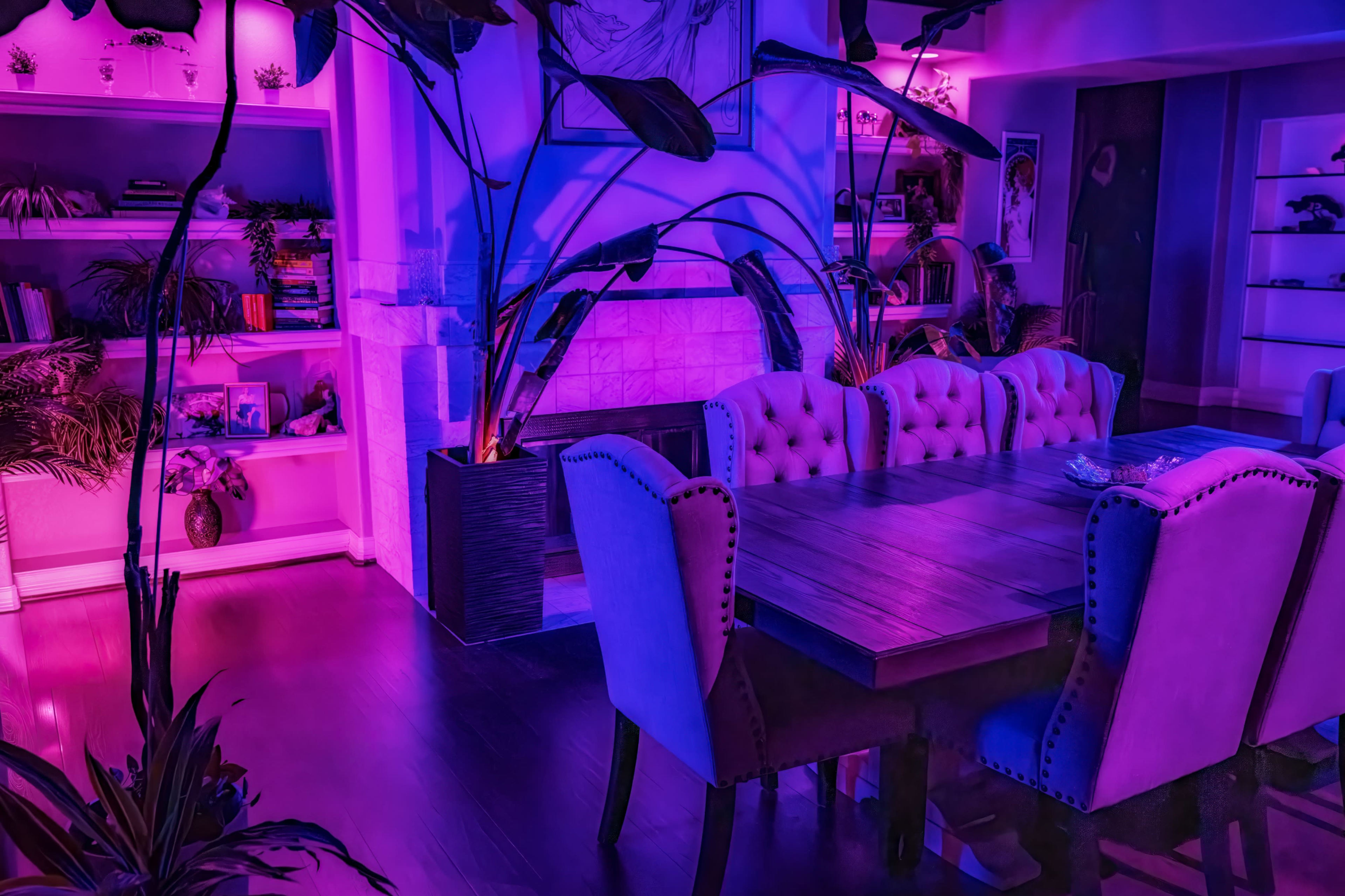 The image shows a dining area with a large wooden table surrounded by upholstered chairs, set against a backdrop of purple lighting and decorative plants.