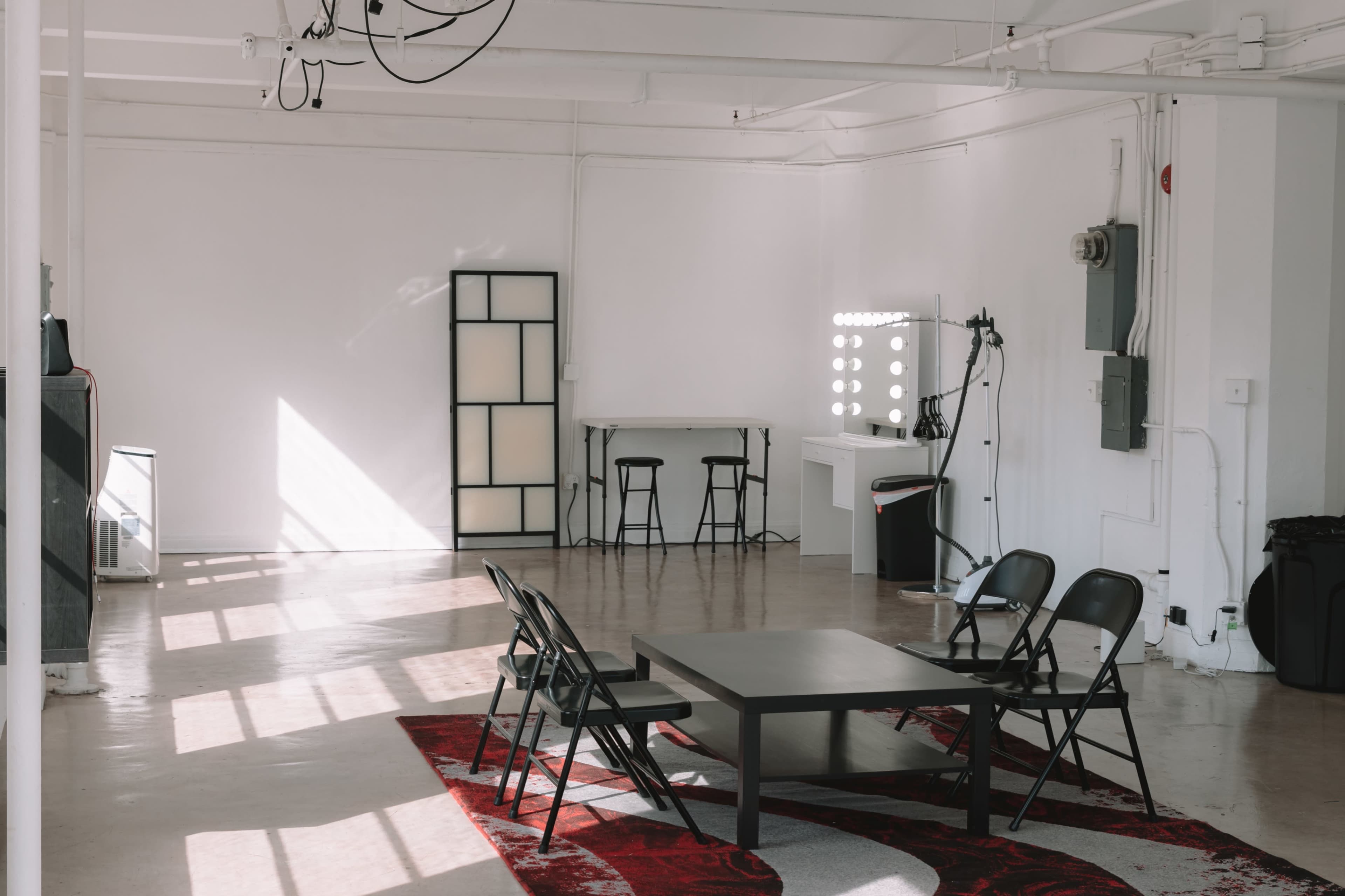 The image shows a minimalist, empty studio space featuring a black table surrounded by folding chairs, a lighted vanity mirror, and large windows allowing natural light in.