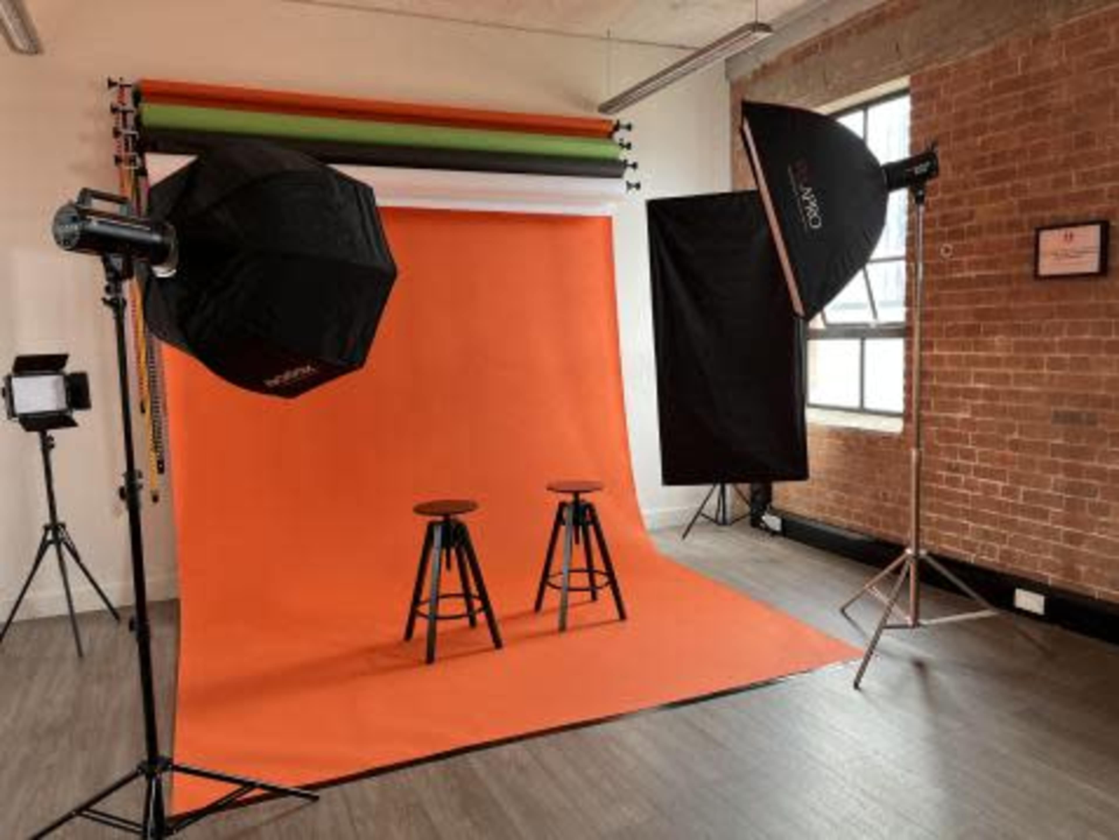 A photography studio setup with an orange backdrop, two black stools, and various lighting equipment.