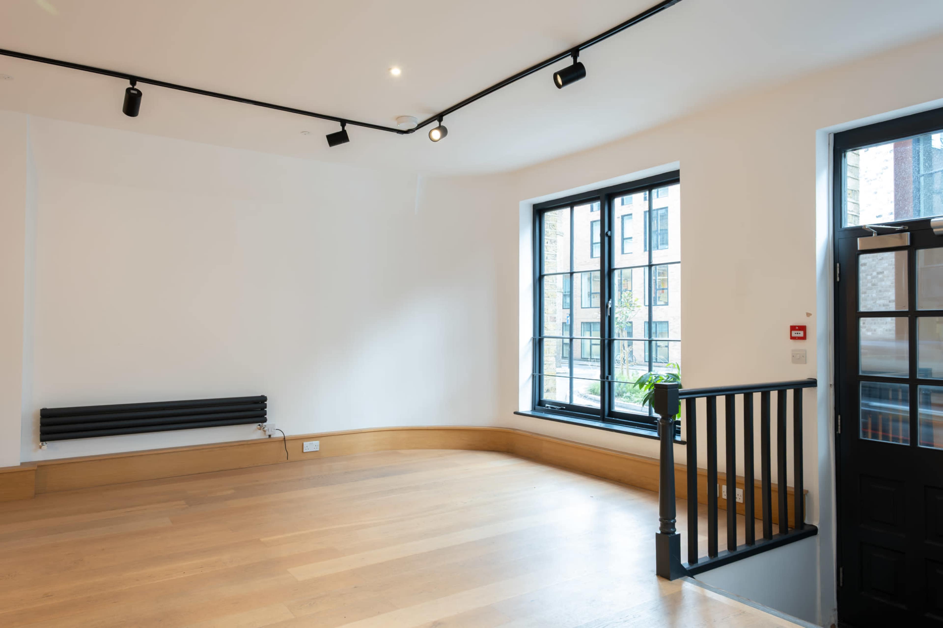 The image shows an empty room with wooden flooring, large windows, and a staircase leading to an upper level.