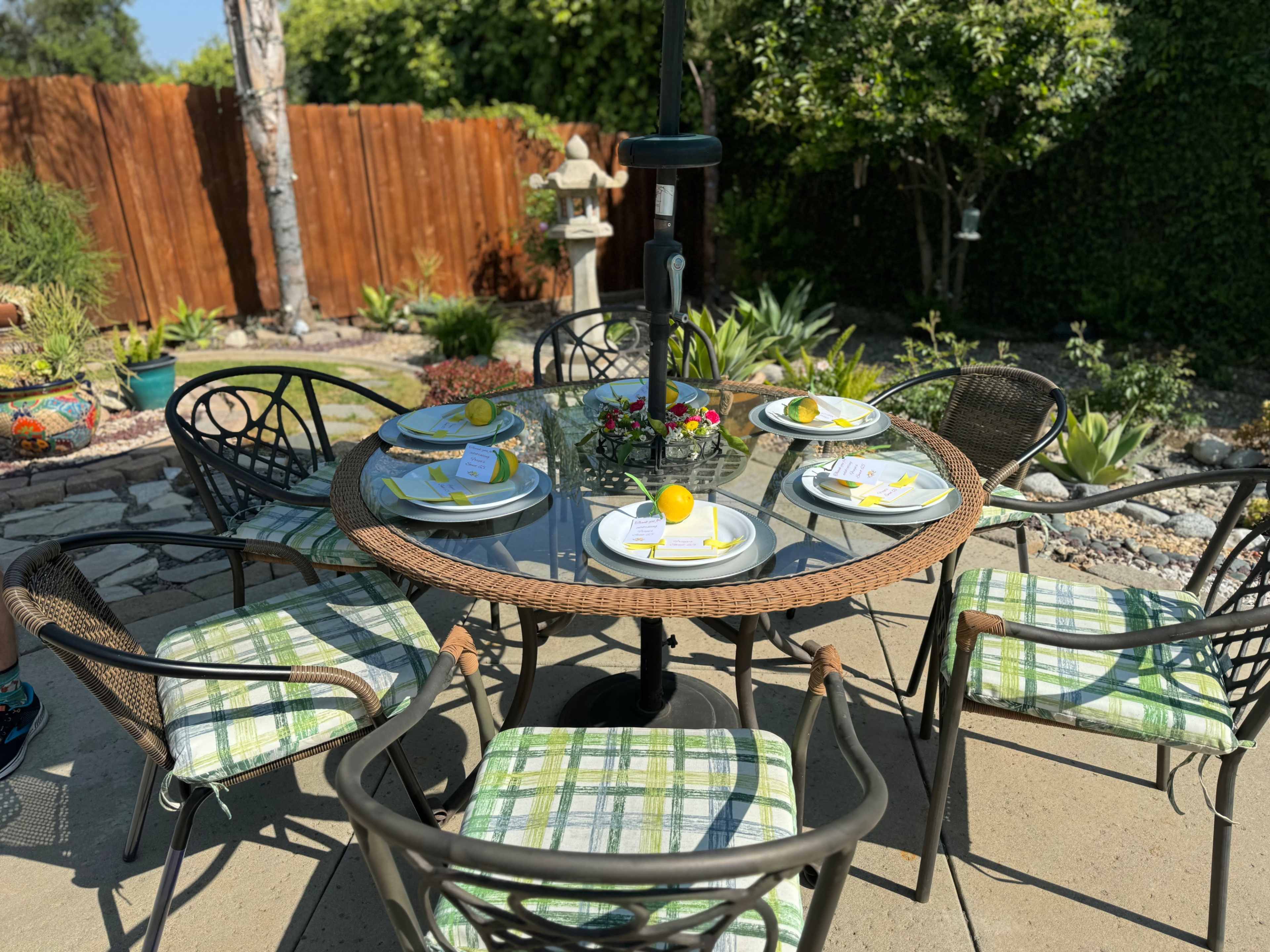 A round table set for six is placed on a patio, surrounded by green plants and a wooden fence.