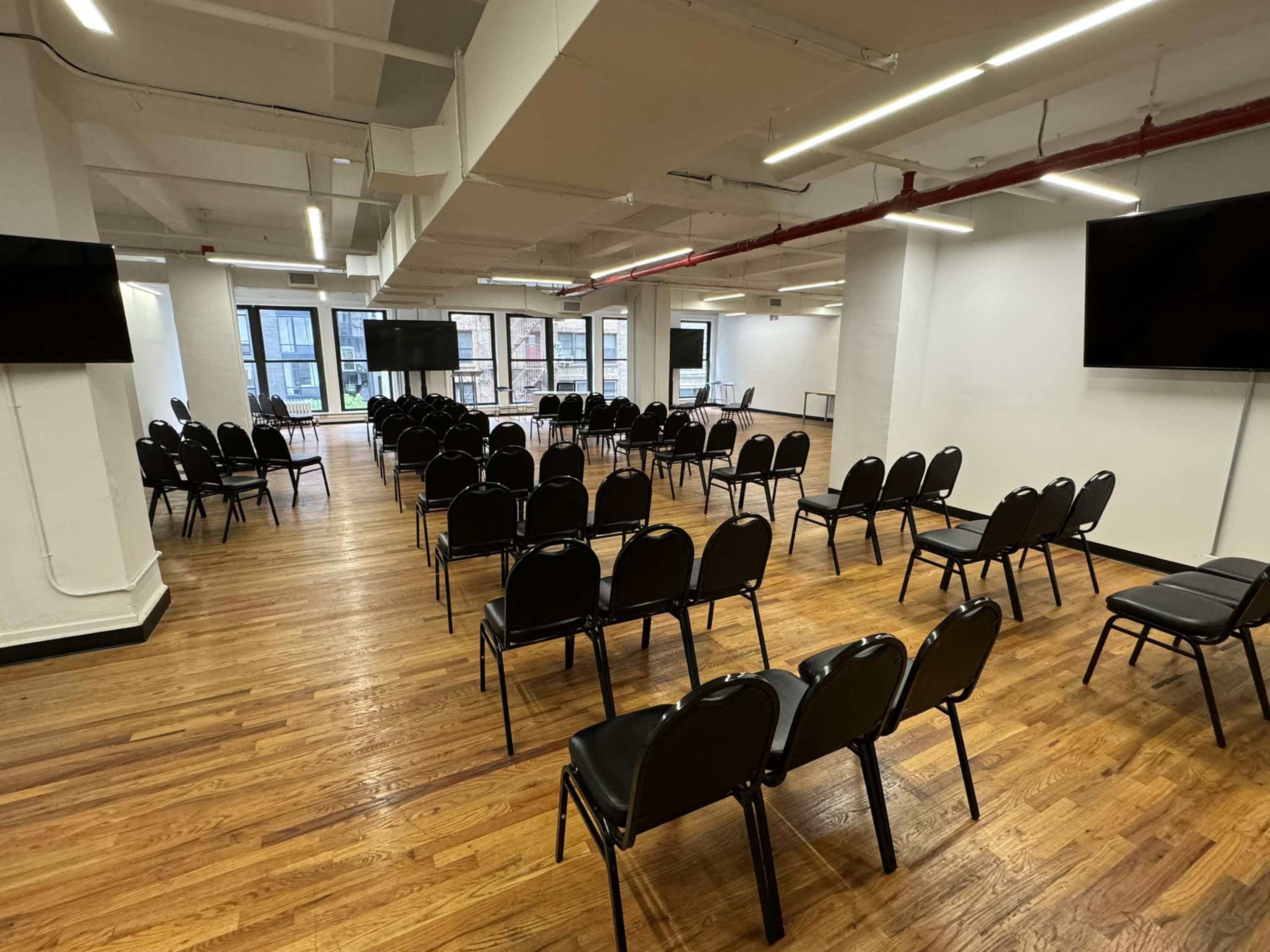 A rows of black chairs are arranged in a large, empty conference room with wooden flooring and large windows.