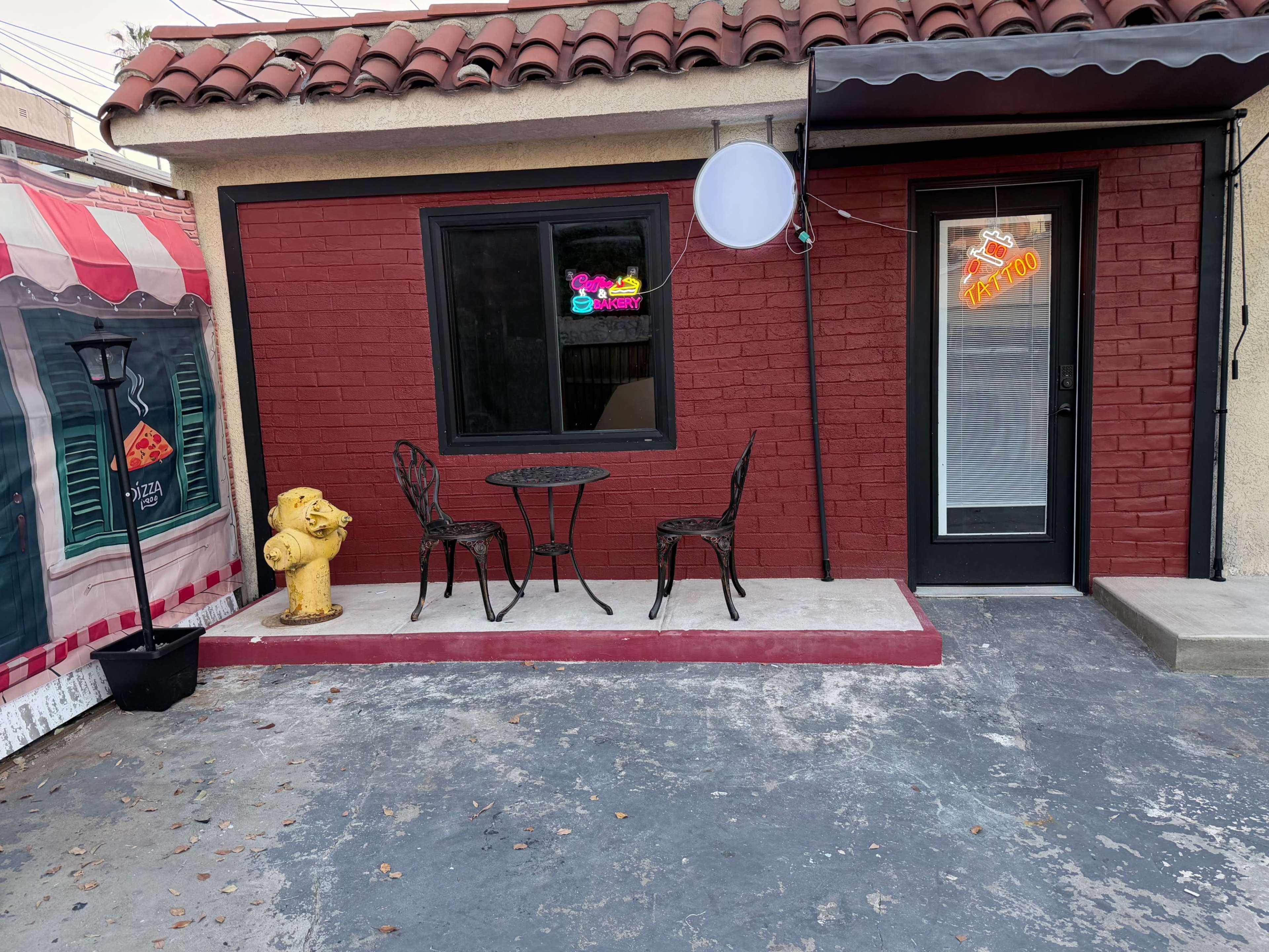 The image shows a small outdoor patio area with a table and two chairs in front of a red brick building, accompanied by a yellow fire hydrant and a hanging neon sign.