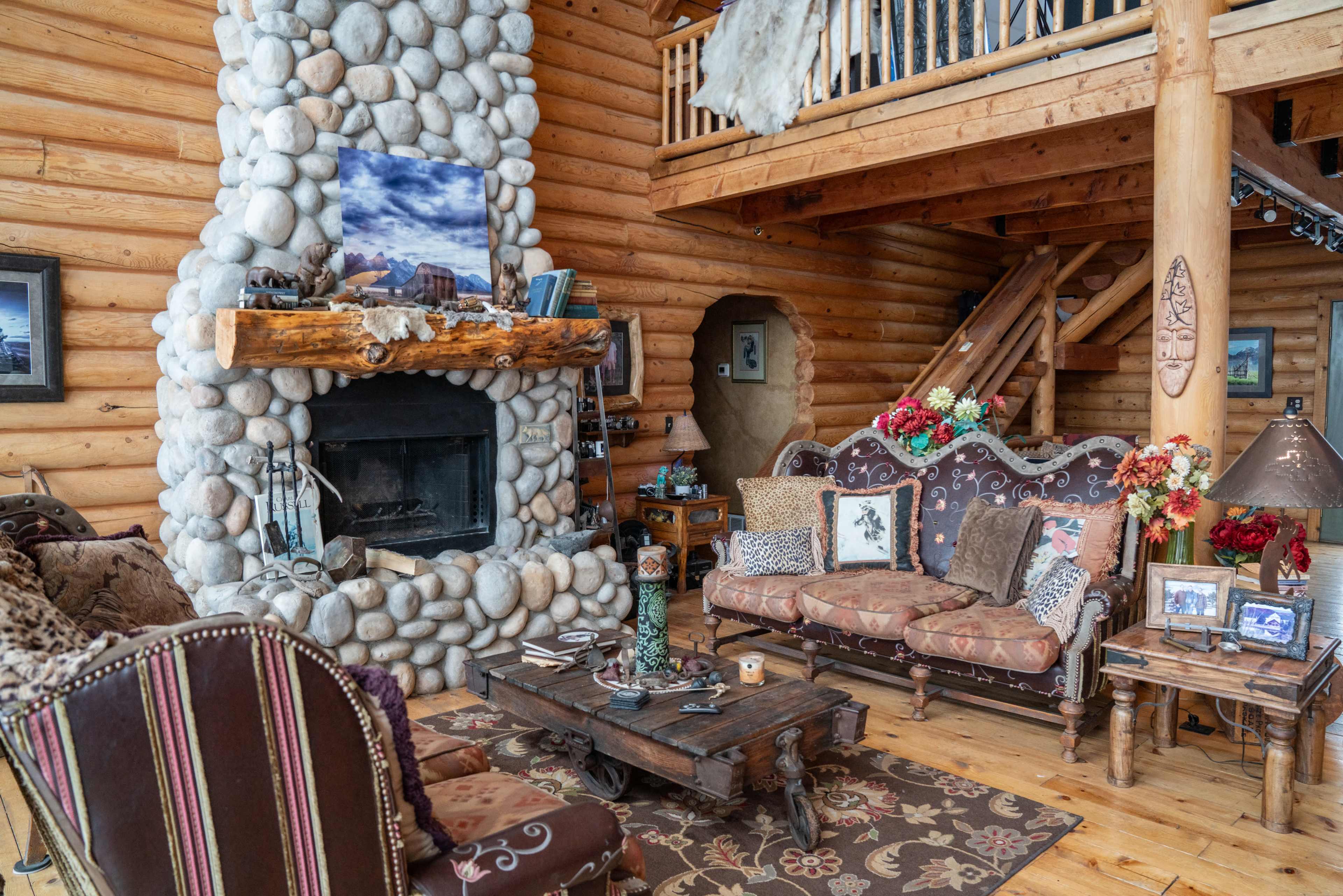 The scene features a rustic log cabin interior with a stone fireplace, wooden beams, and vintage furniture, including a sofa and armchairs adorned with floral decorations.