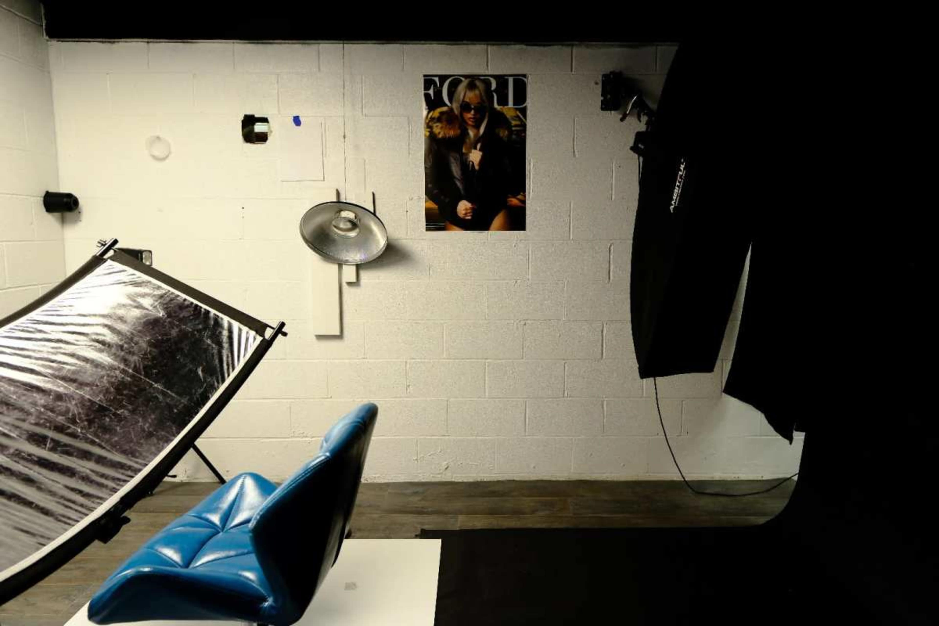 The image shows a photography studio featuring a blue chair, a reflector, a lighting fixture, and a fashion magazine poster on the wall.
