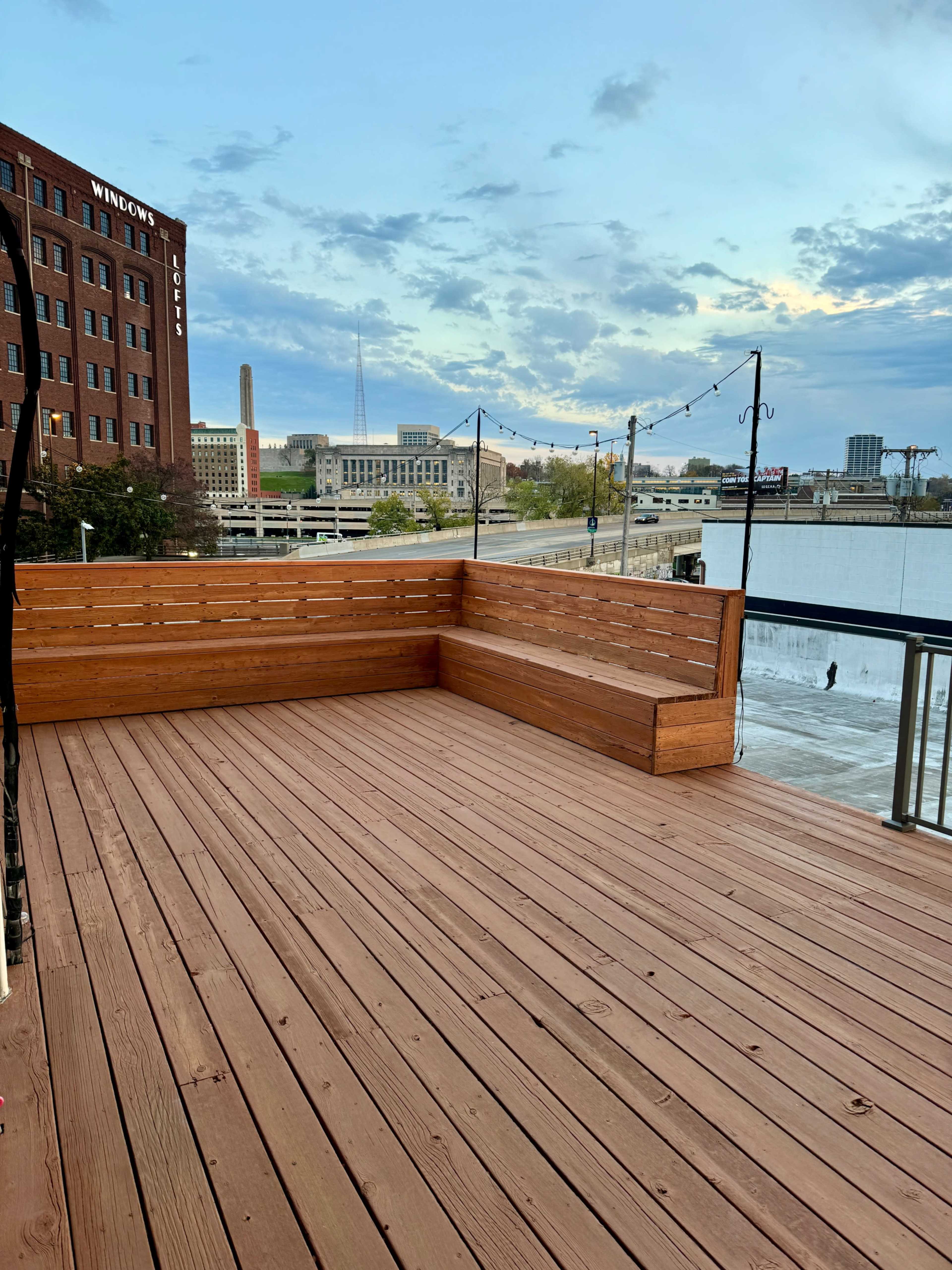 The image shows a wooden deck with a bench, overlooking a river and nearby buildings under a cloudy sky.
