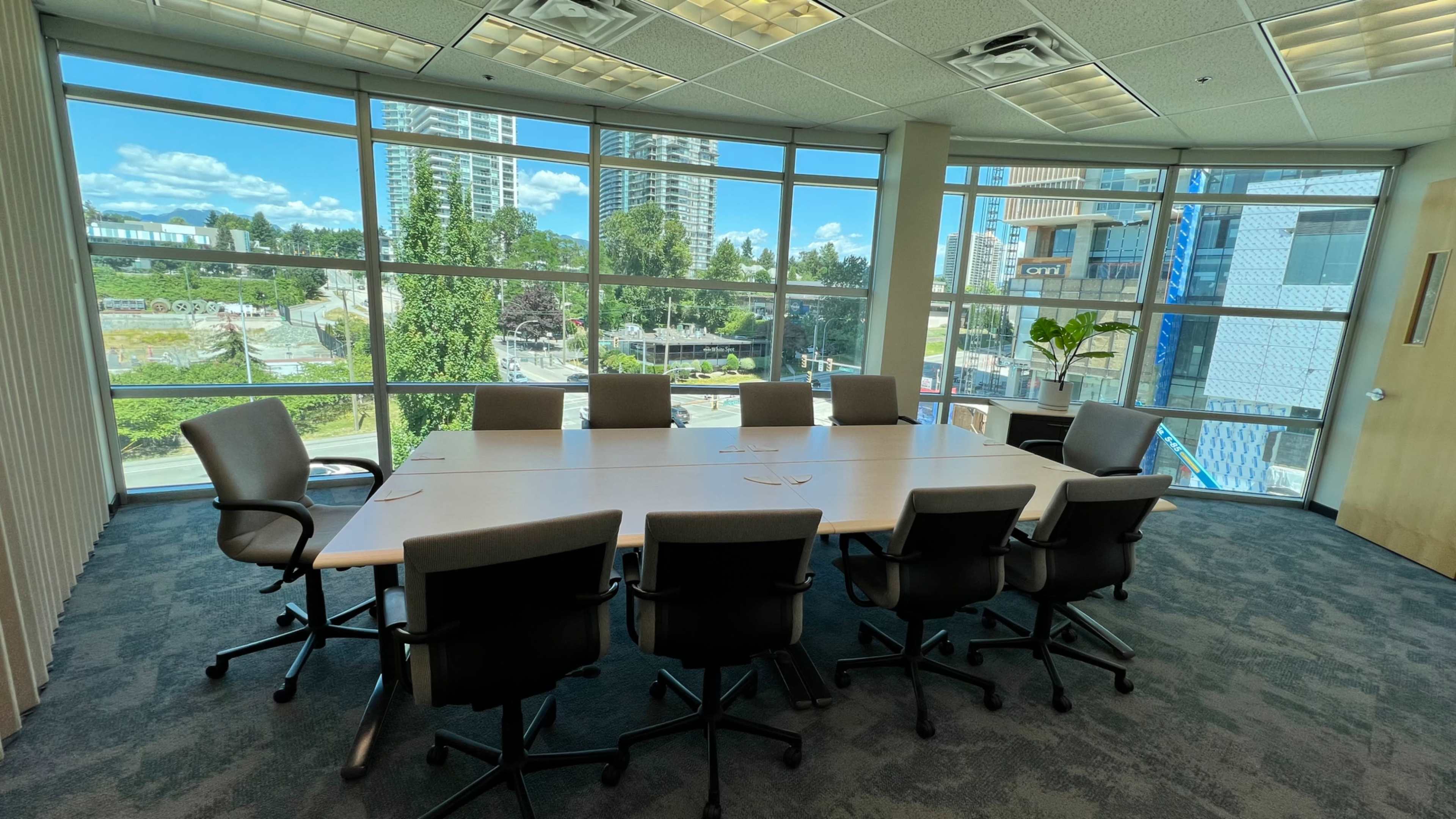 A boardroom with a long table and eight chairs is set against large windows offering a view of nearby buildings and green spaces.