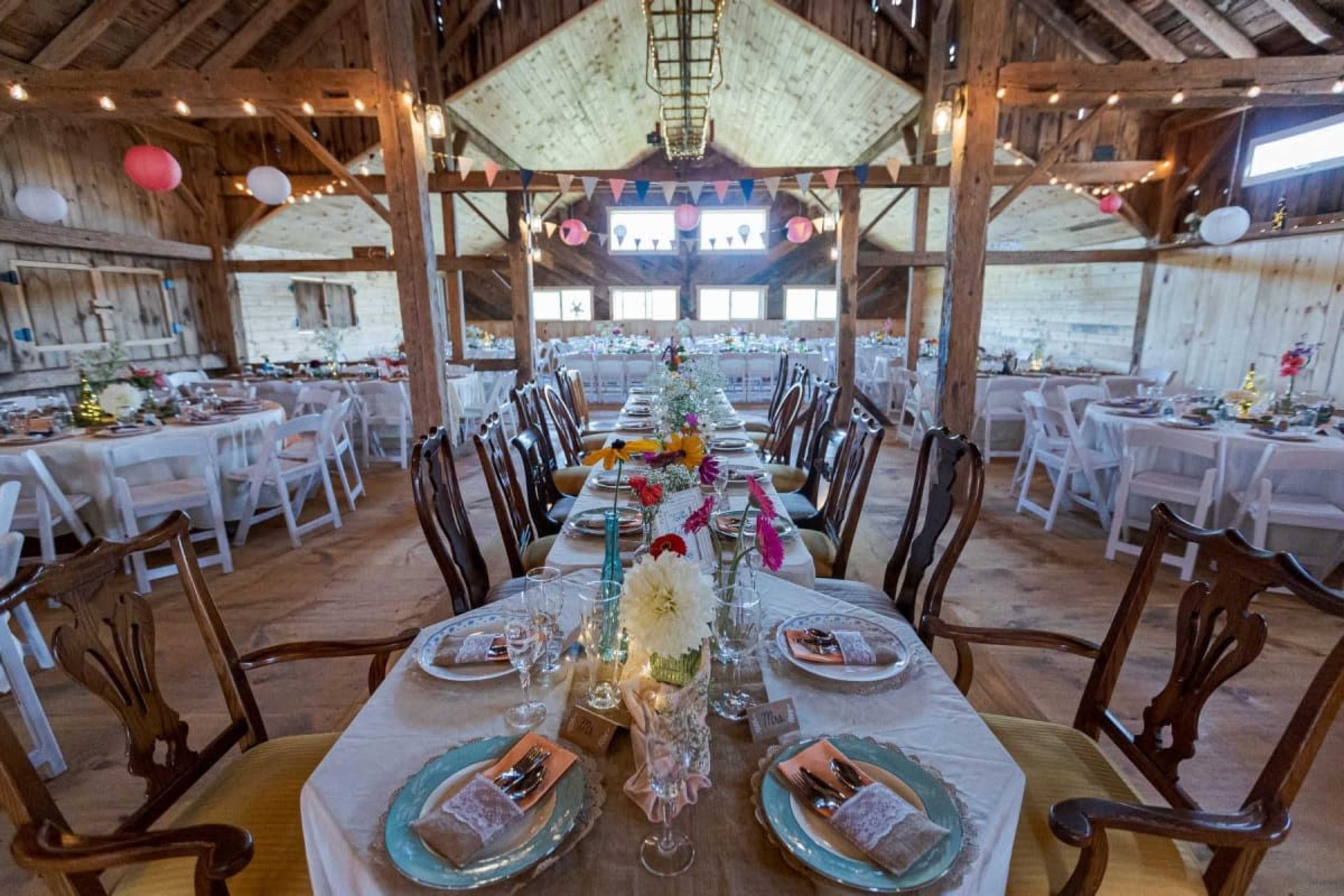 The image shows a spacious barn interior set for a dining event, featuring long tables adorned with floral centerpieces and decorative table settings.