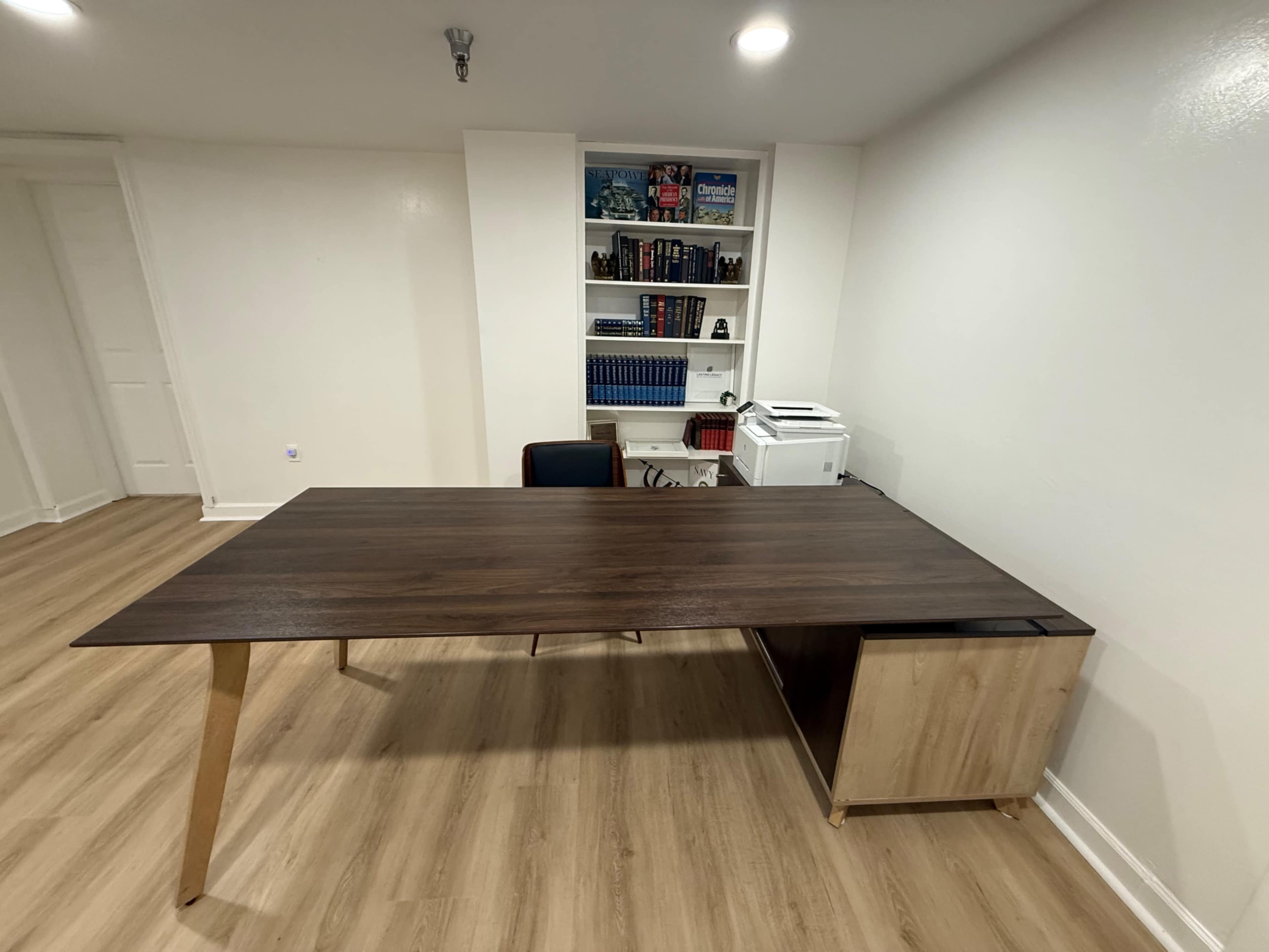 The image shows a modern office space featuring a wooden desk with a chair and a shelf filled with books in the background.