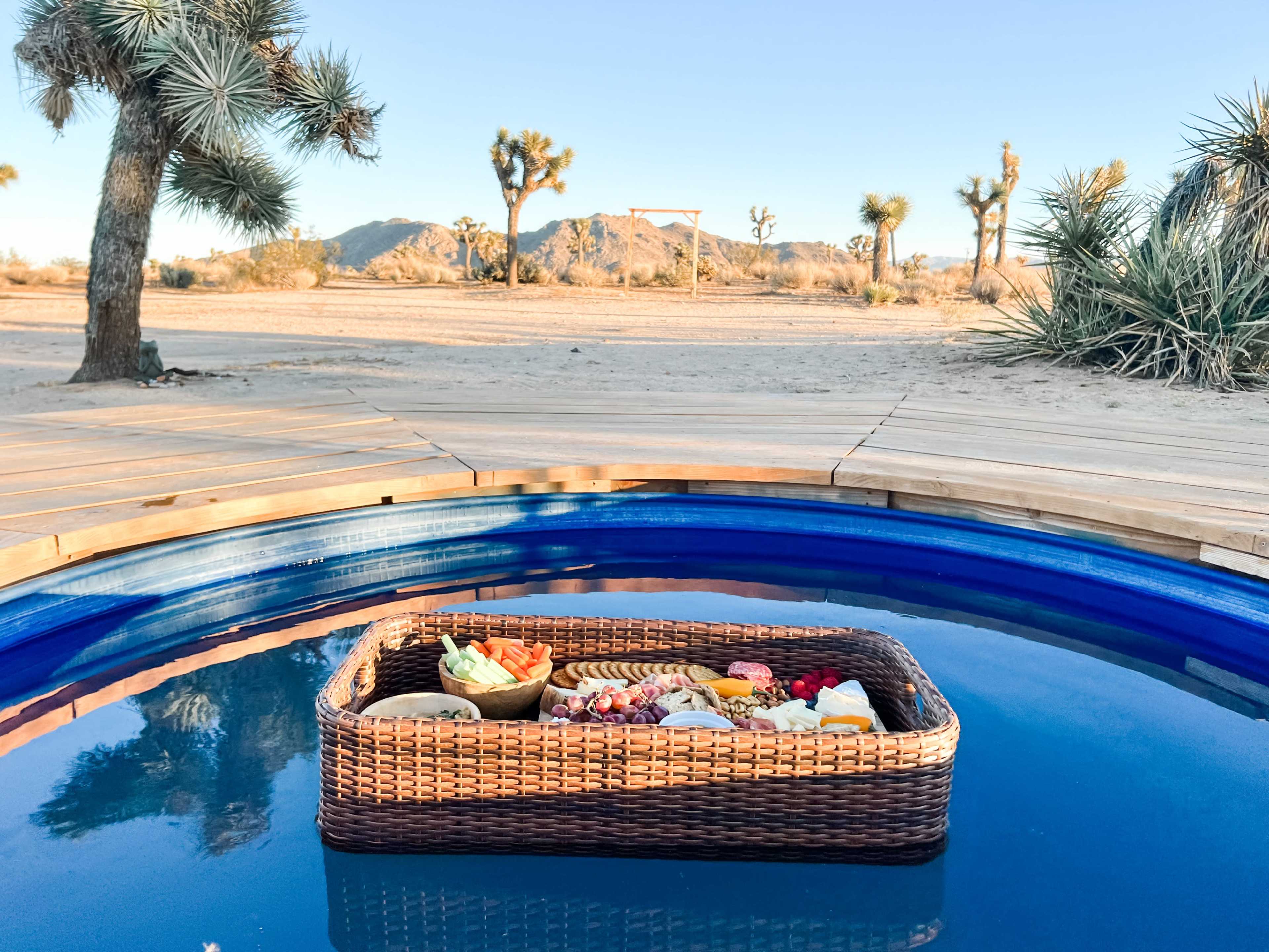A wicker basket containing an assortment of fruits and snacks floats in a blue pool, set against a desert backdrop with Joshua trees and distant mountains.