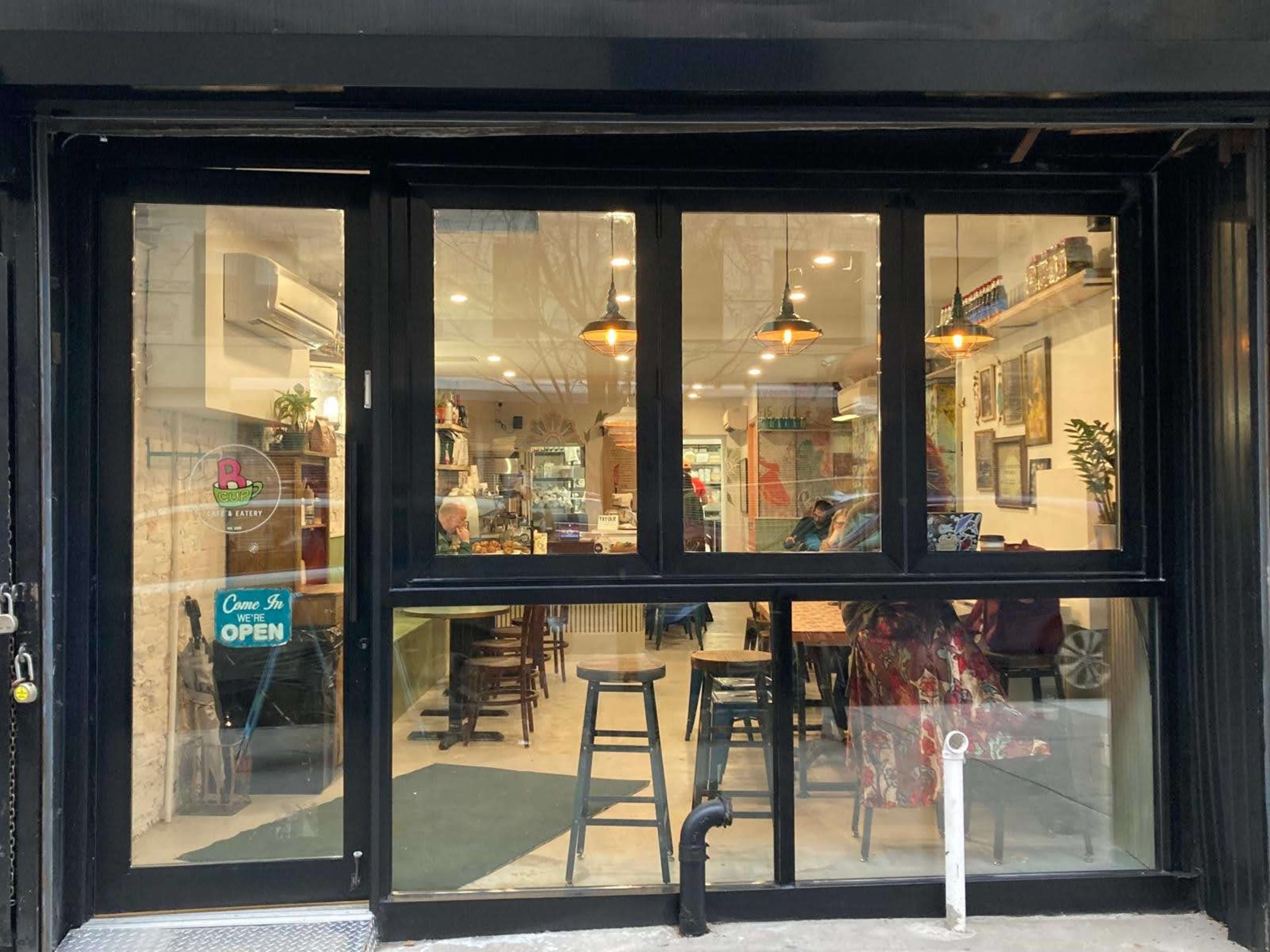 The image shows a cafe with large glass windows, showcasing interior seating and decor, and a sign indicating that it is open.