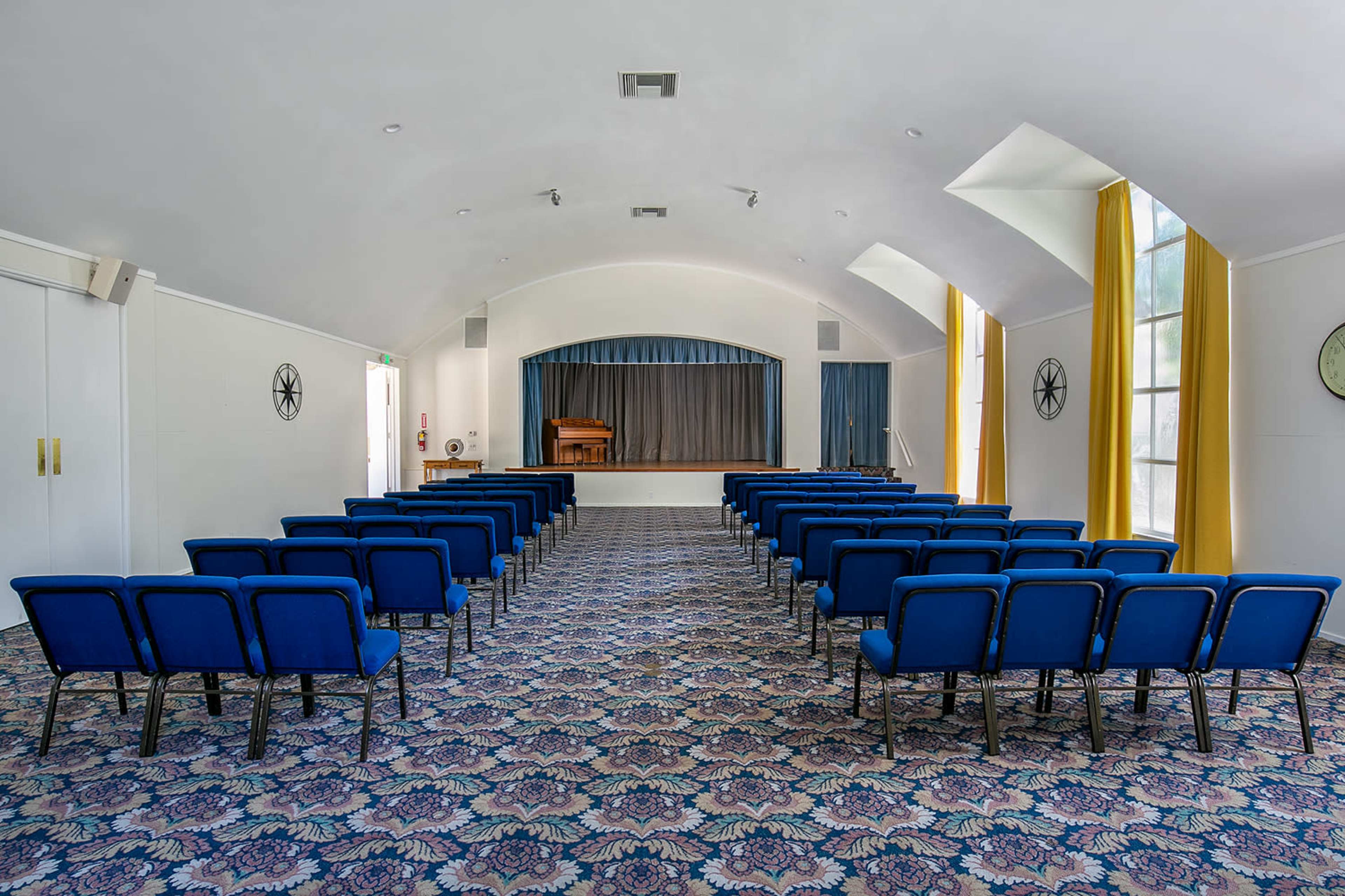 The image shows a large, empty auditorium with blue chairs arranged in rows facing a stage, flanked by yellow curtains.