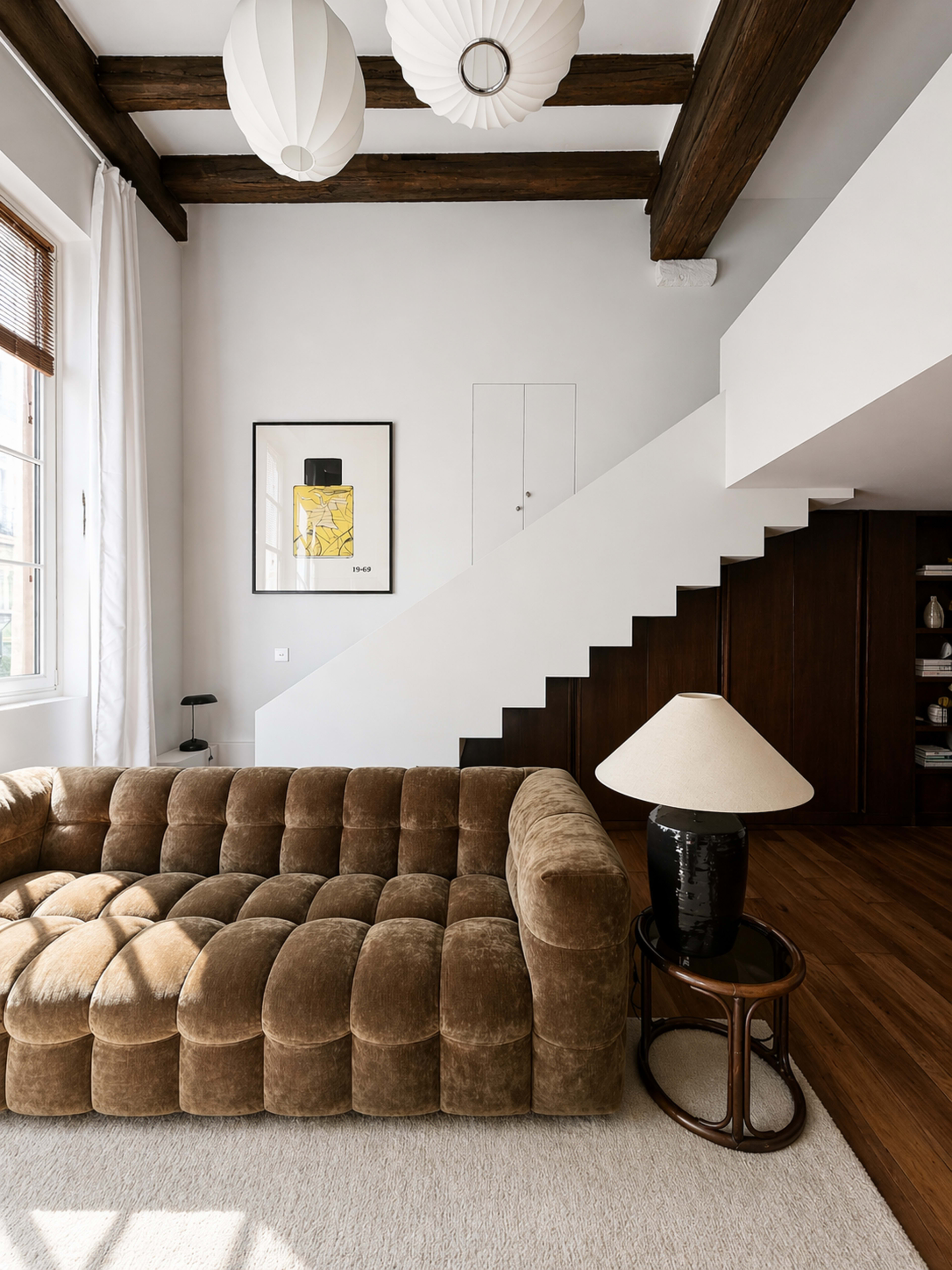The image features a modern living room with a brown tufted sofa, a round side table, and a lamp, set against white walls and wooden beams, with a staircase in the background.