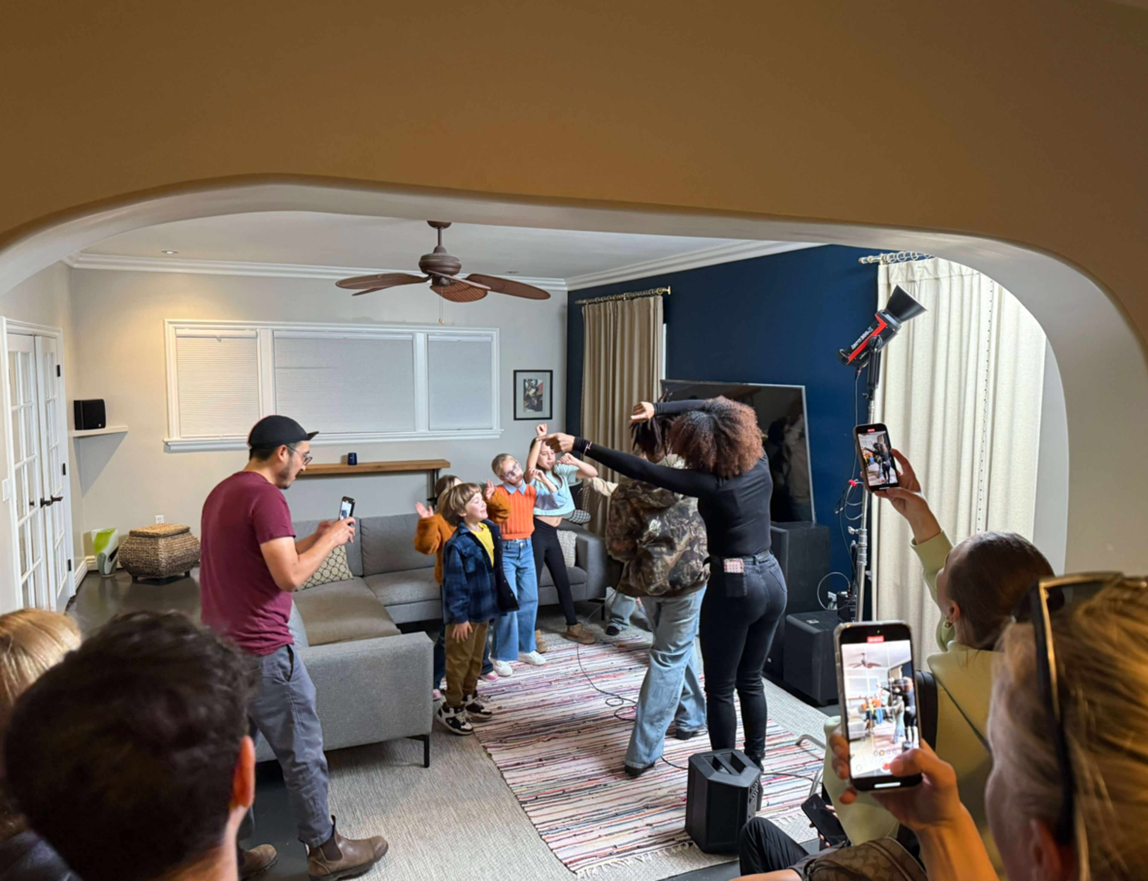 A group of children and adults are gathered in a living room, with some people recording on their phones while others are participating in a musical performance.