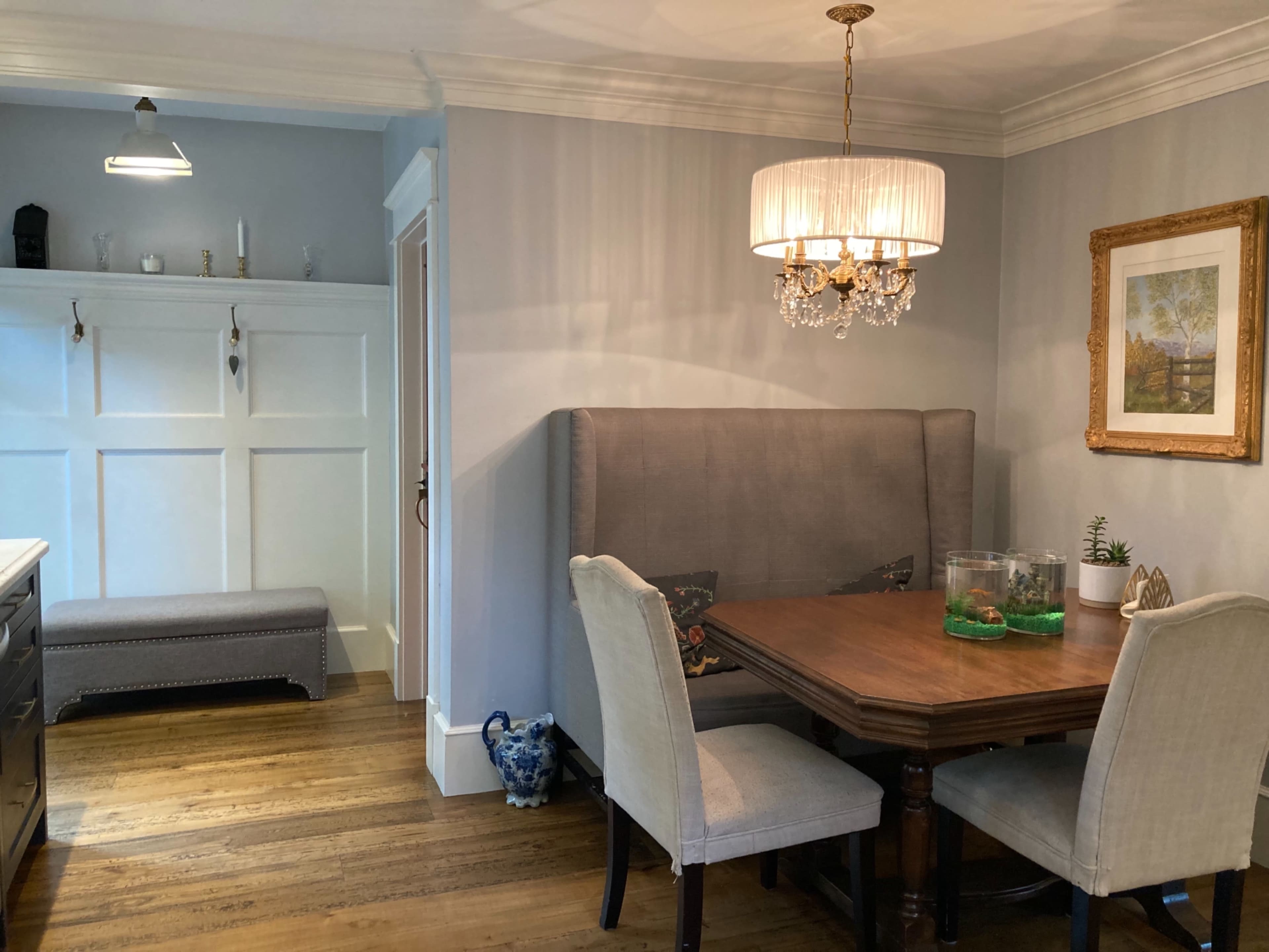 A dining area featuring a wooden table with upholstered chairs, a clear fish tank on the table, and a cozy nook with a bench against the wall, all illuminated by a chandelier.