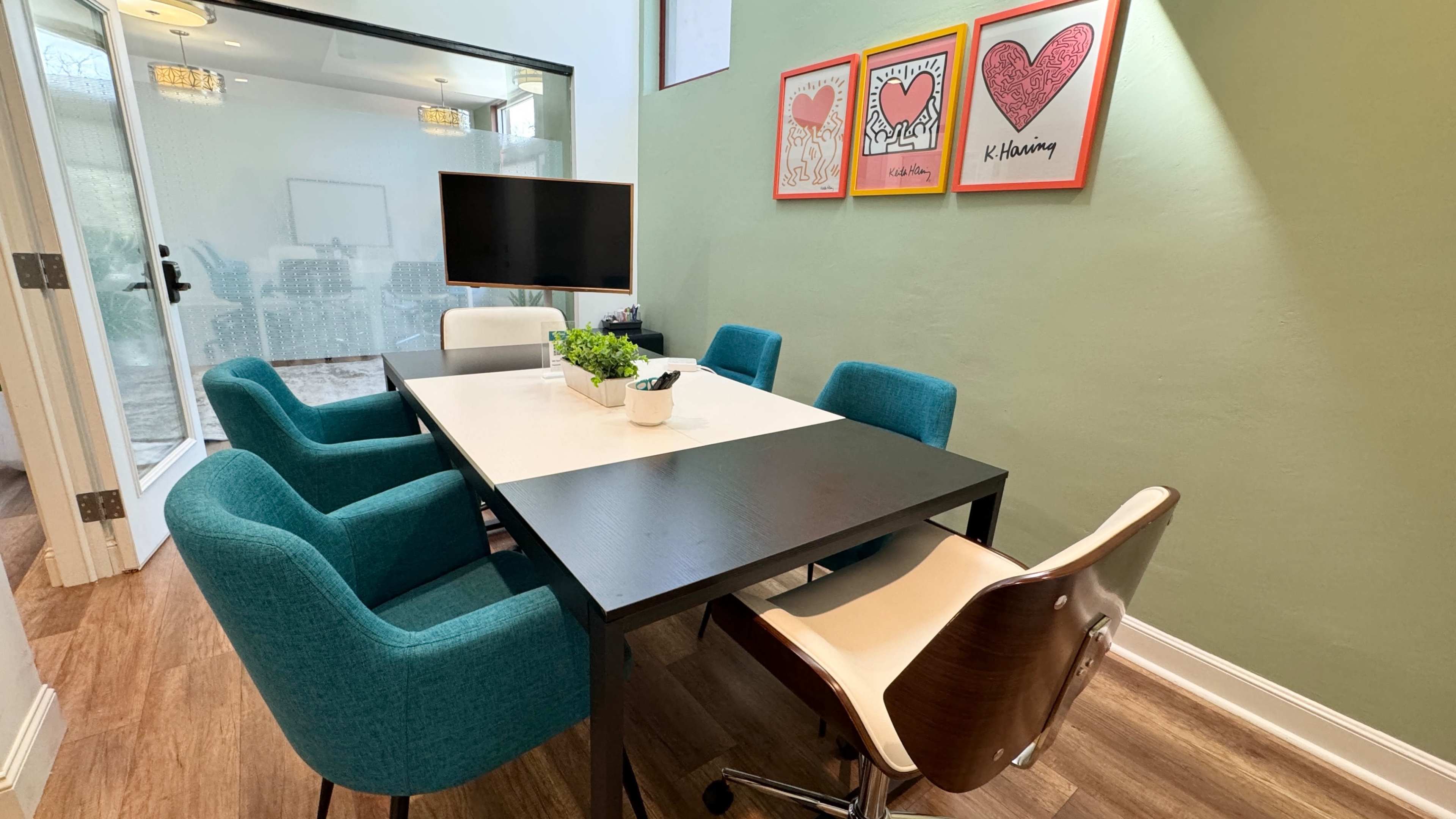 A modern meeting room with a rectangular black table surrounded by six chairs, three in teal and two in cream, alongside a television and framed heart artwork on the wall.