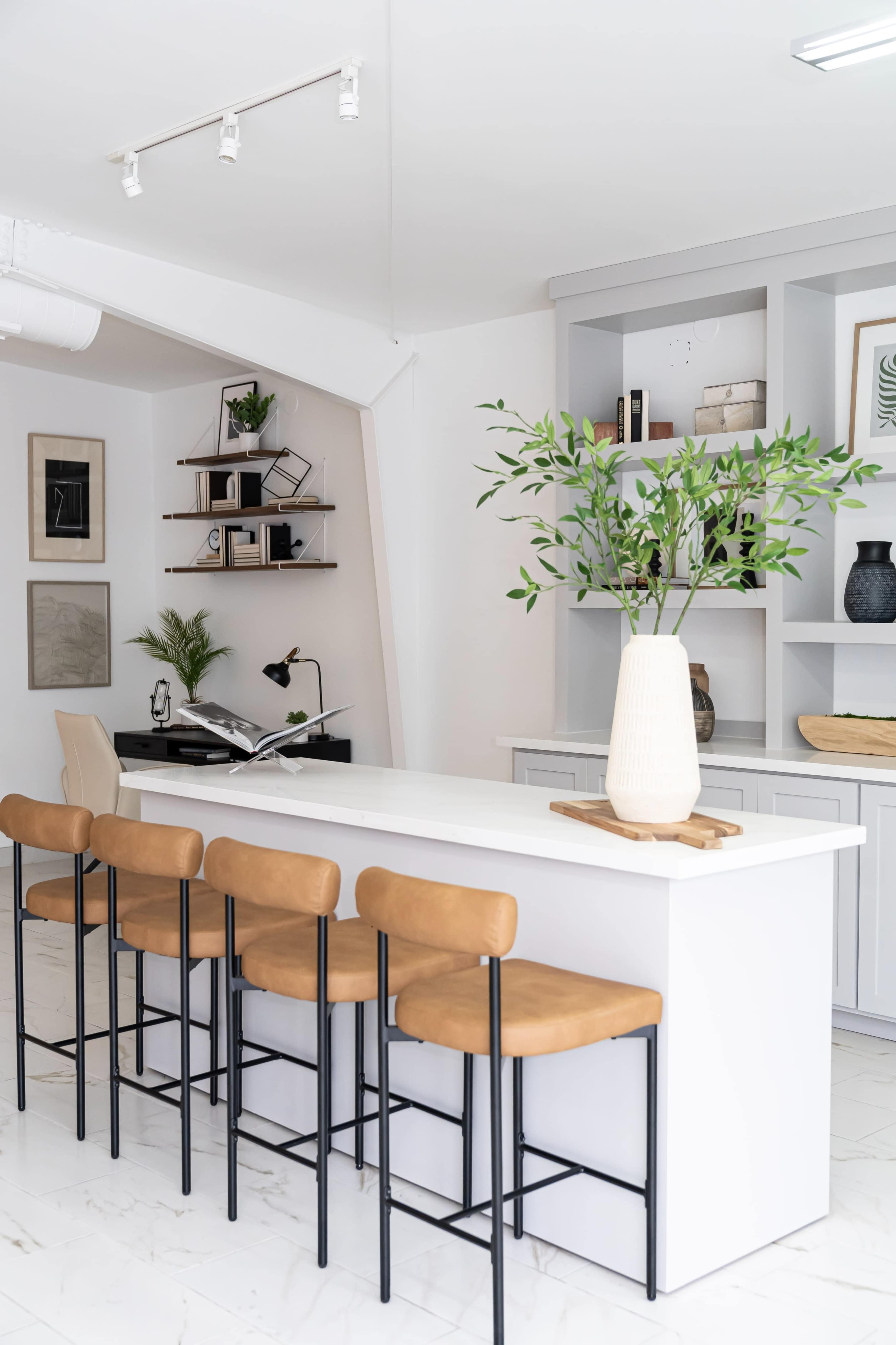 A modern kitchen features a white island with six tan, high-backed stools and a shelf holding decorative items in the background.