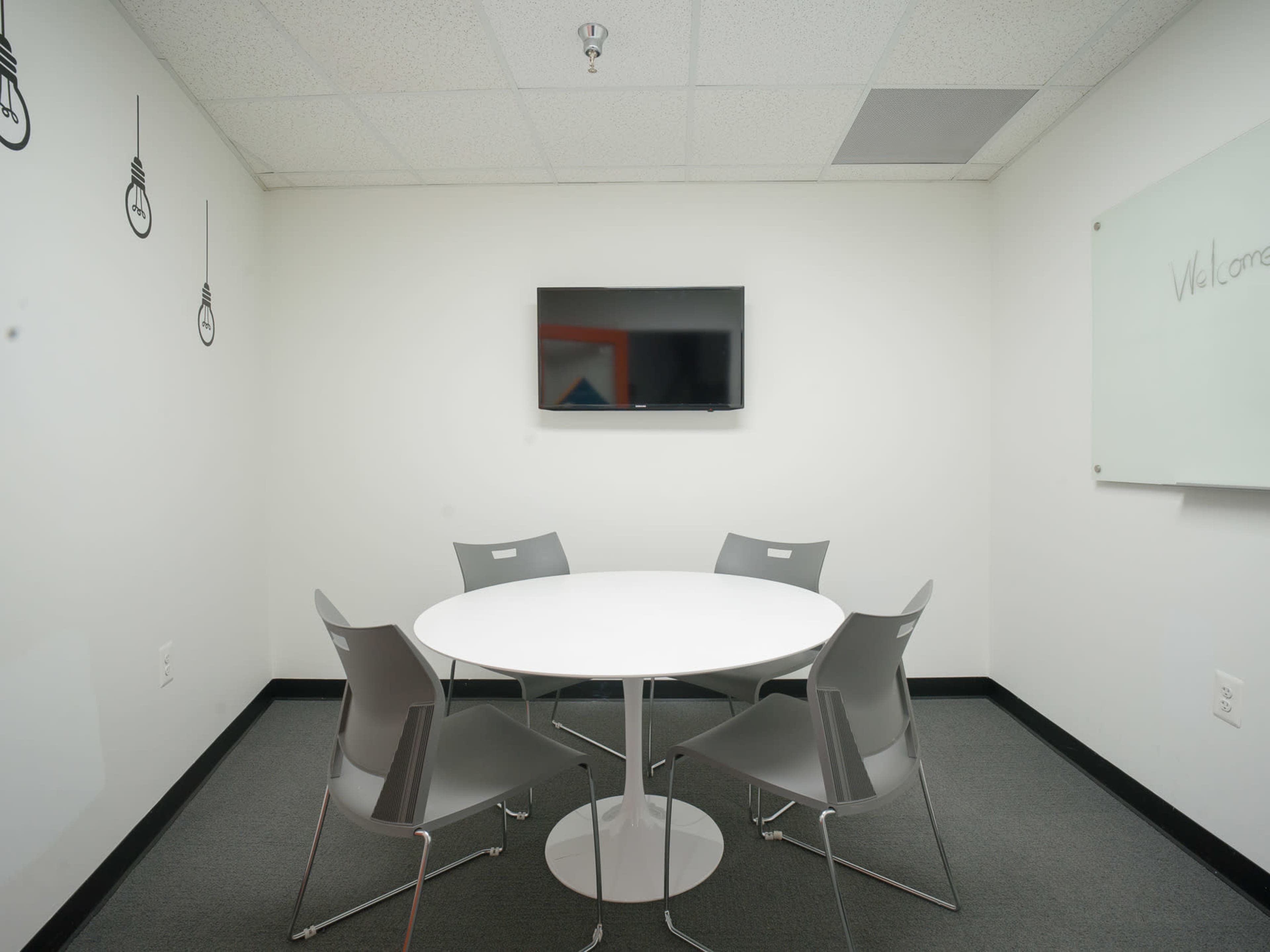 A small meeting room features a round table surrounded by five chairs, complemented by a wall-mounted screen.