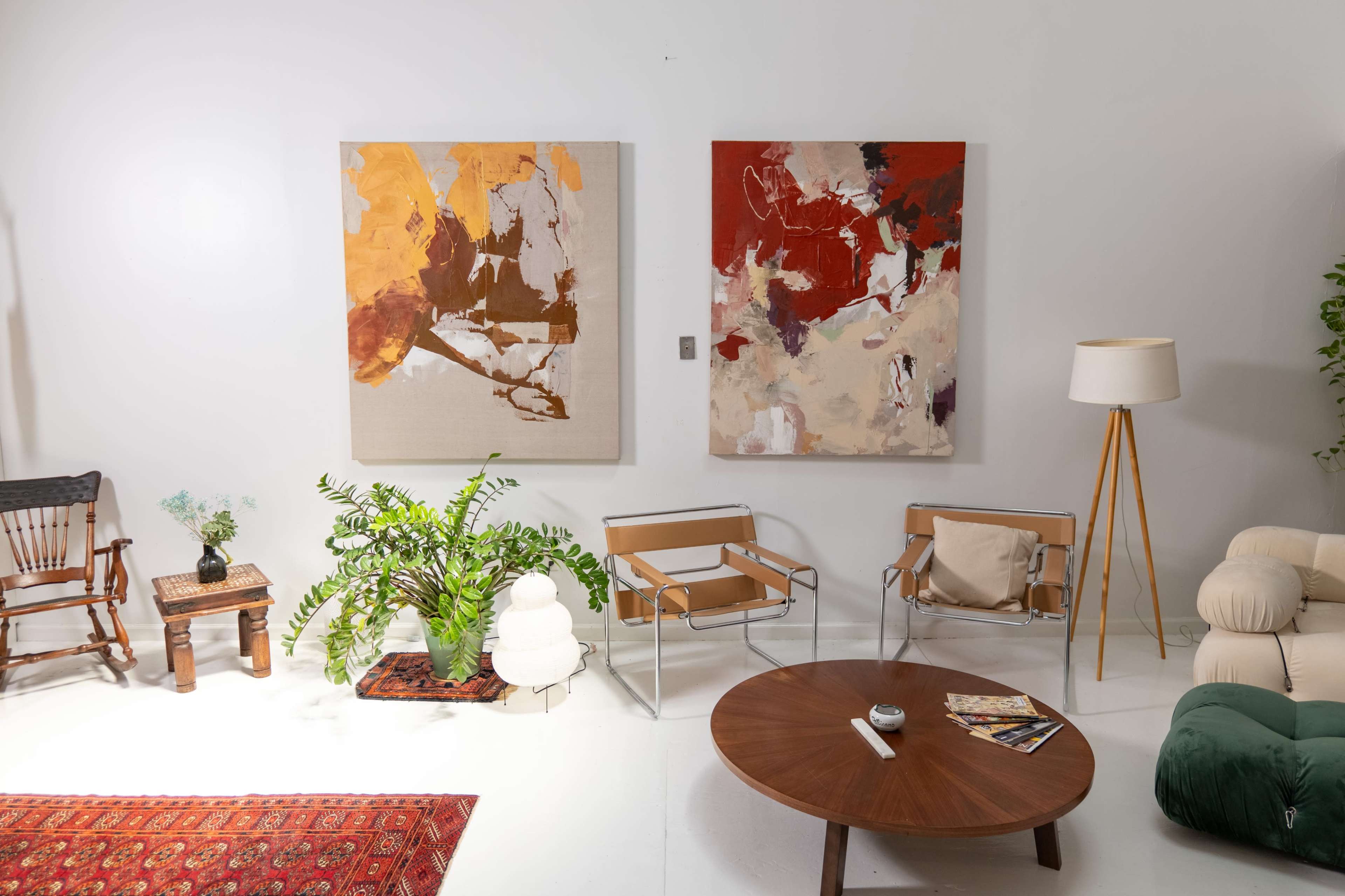 The scene features a modern living area with two abstract paintings on the walls, a round wooden coffee table at the center, stylish chairs, and greenery.