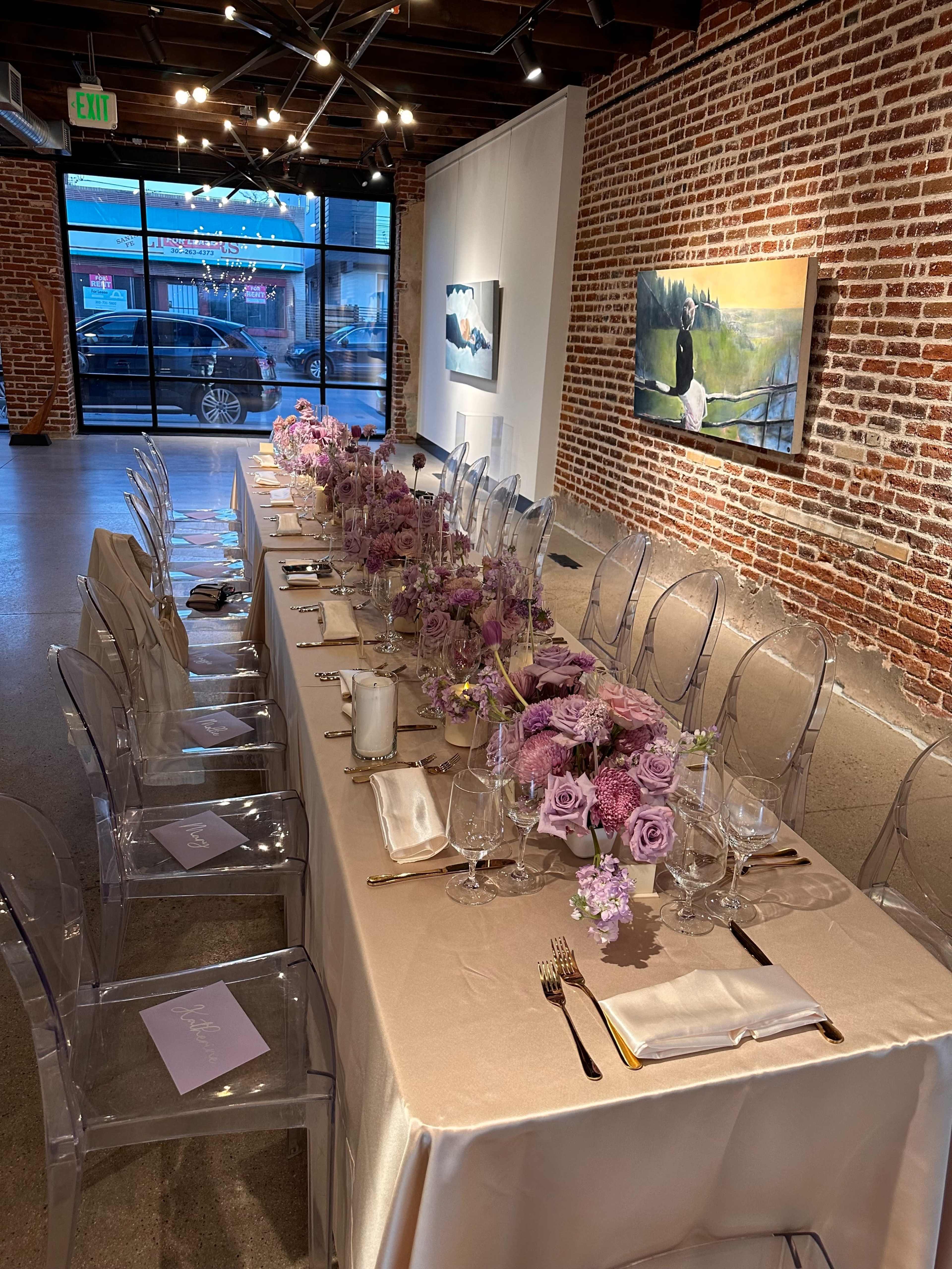 A long table is set for an event, adorned with lavender flowers and elegant tableware, in a brick-walled gallery space.