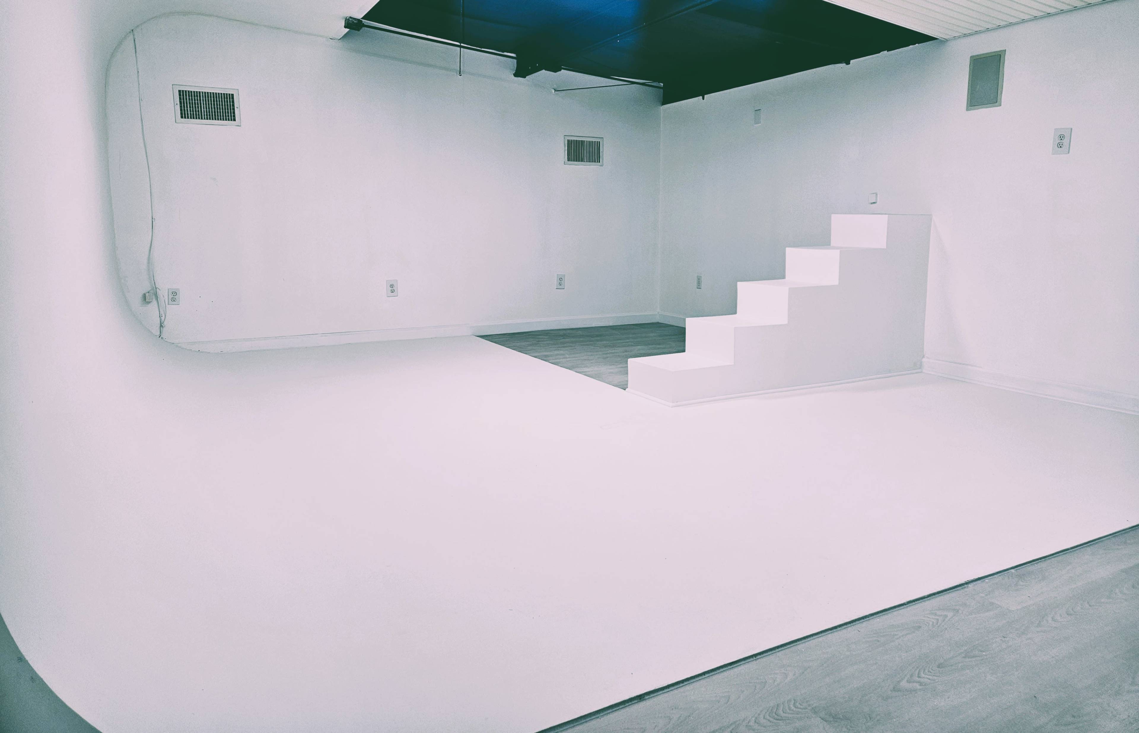 The image shows a minimalist, empty room featuring a curved white wall and a set of white steps leading to a raised platform.
