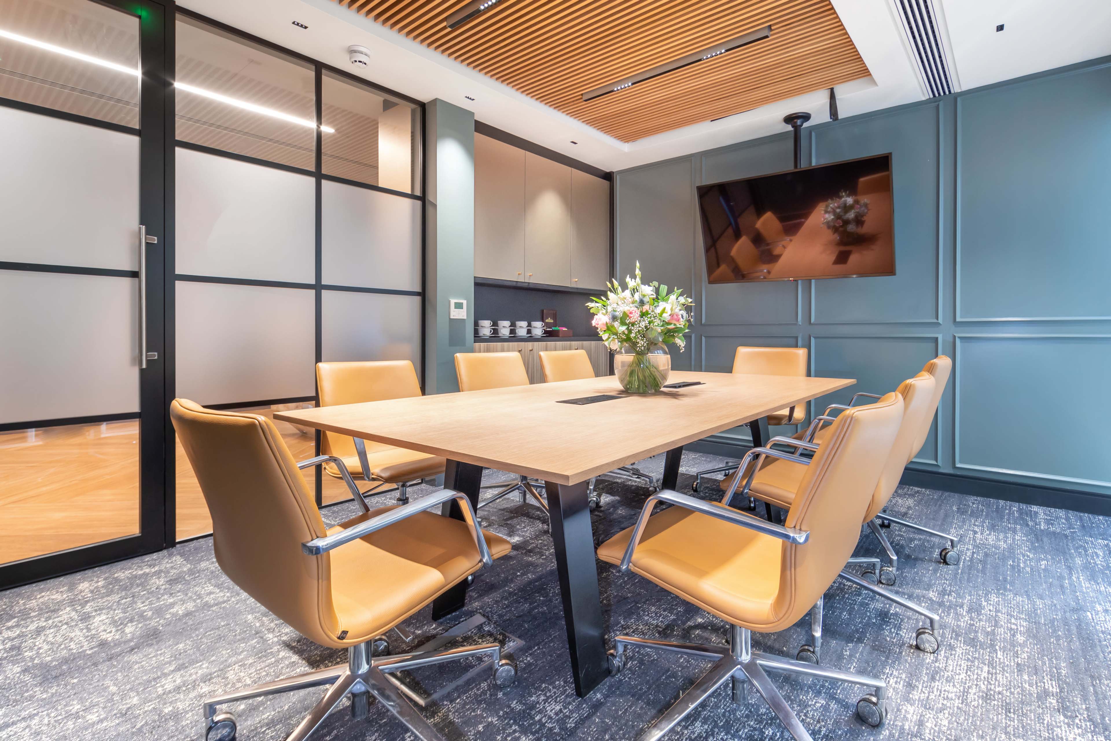 A modern conference room features a large wooden table surrounded by eight leather chairs, with a flat-screen TV mounted on the wall and a floral arrangement on the table.