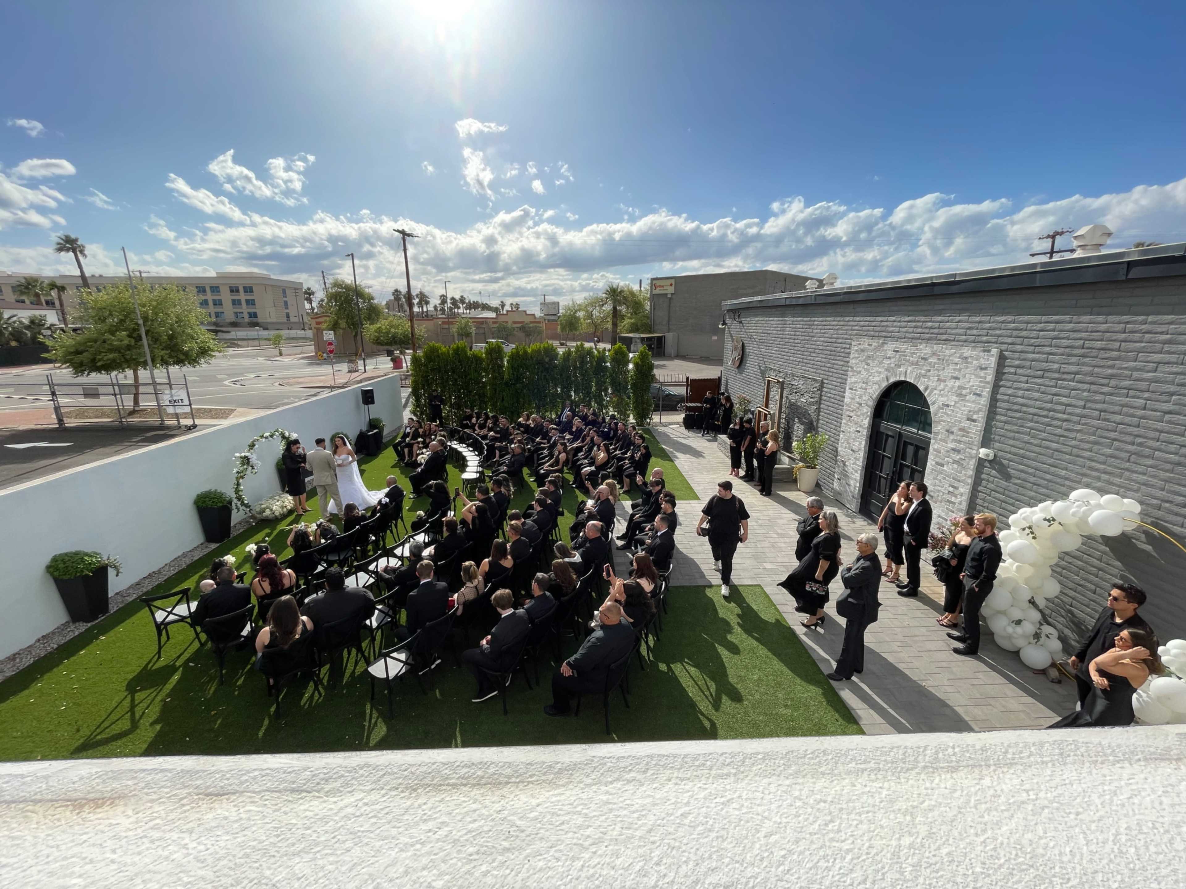 A wedding ceremony takes place outdoors with numerous seated guests facing a couple standing at the front, under a blue sky with scattered clouds.