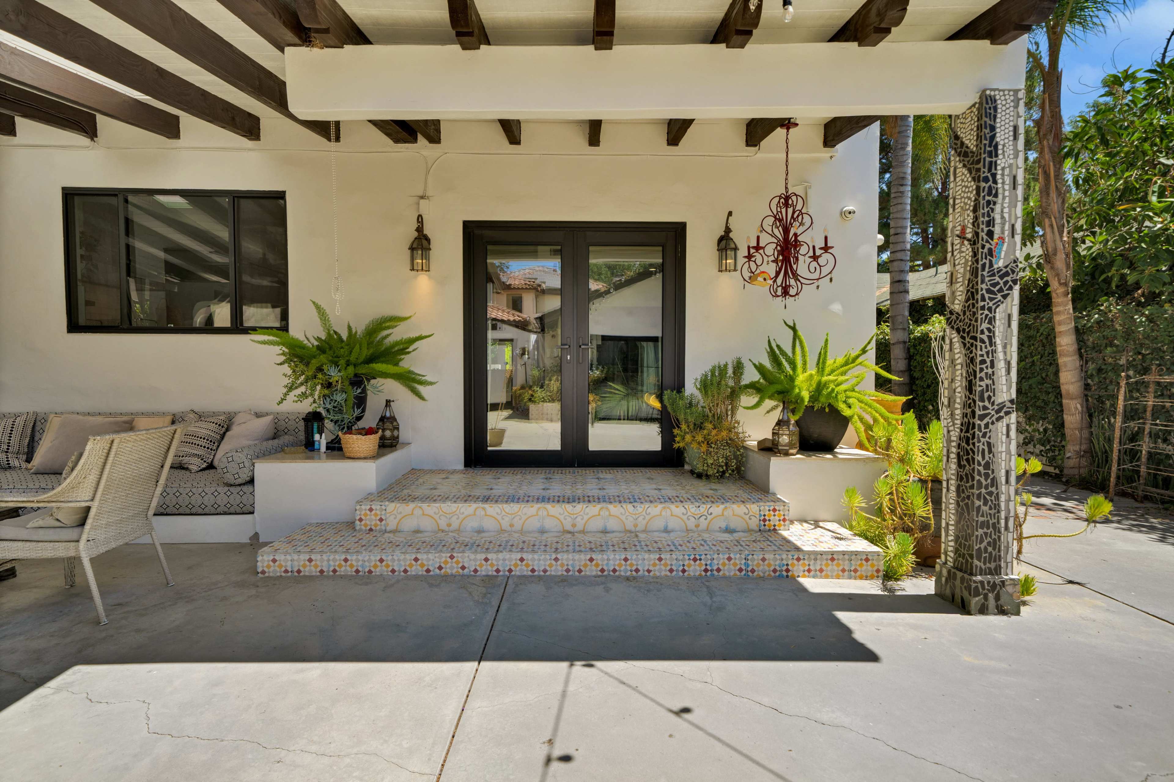 The image shows a covered patio area with steps leading up to double black doors, surrounded by potted plants and decorative tiles.