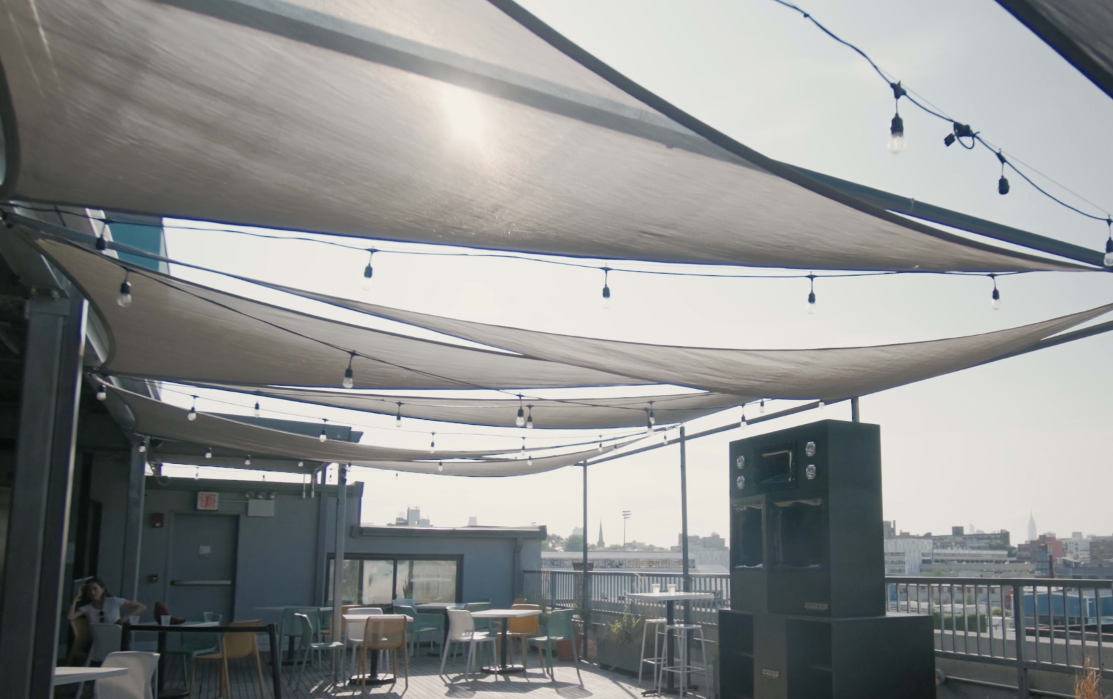 The image shows a rooftop space with overhead fabric canopies, string lights, and scattered tables and chairs.