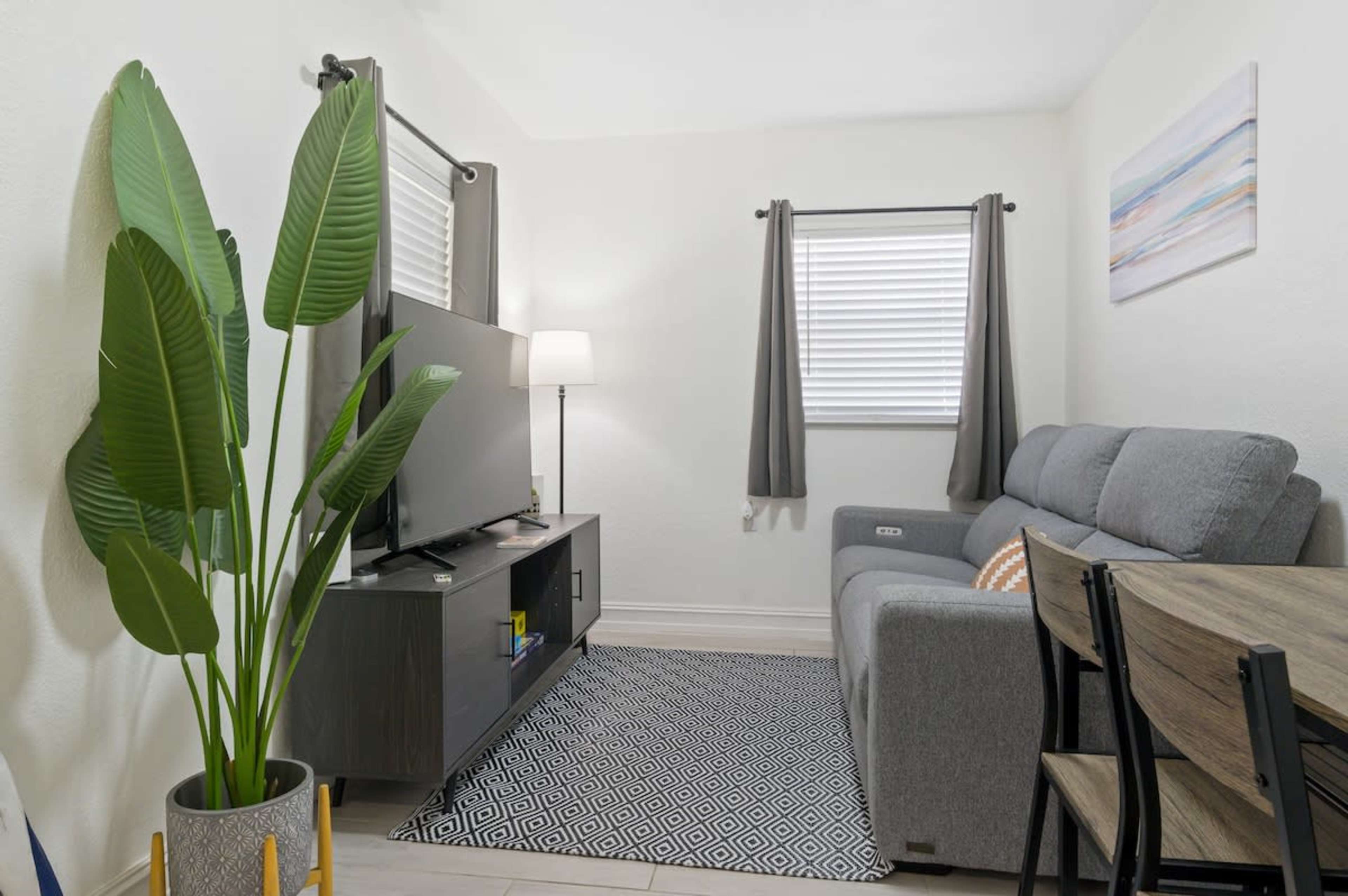 The image shows a small living room featuring a gray sofa, a television on a black media console, a dining table, and a tall potted plant.