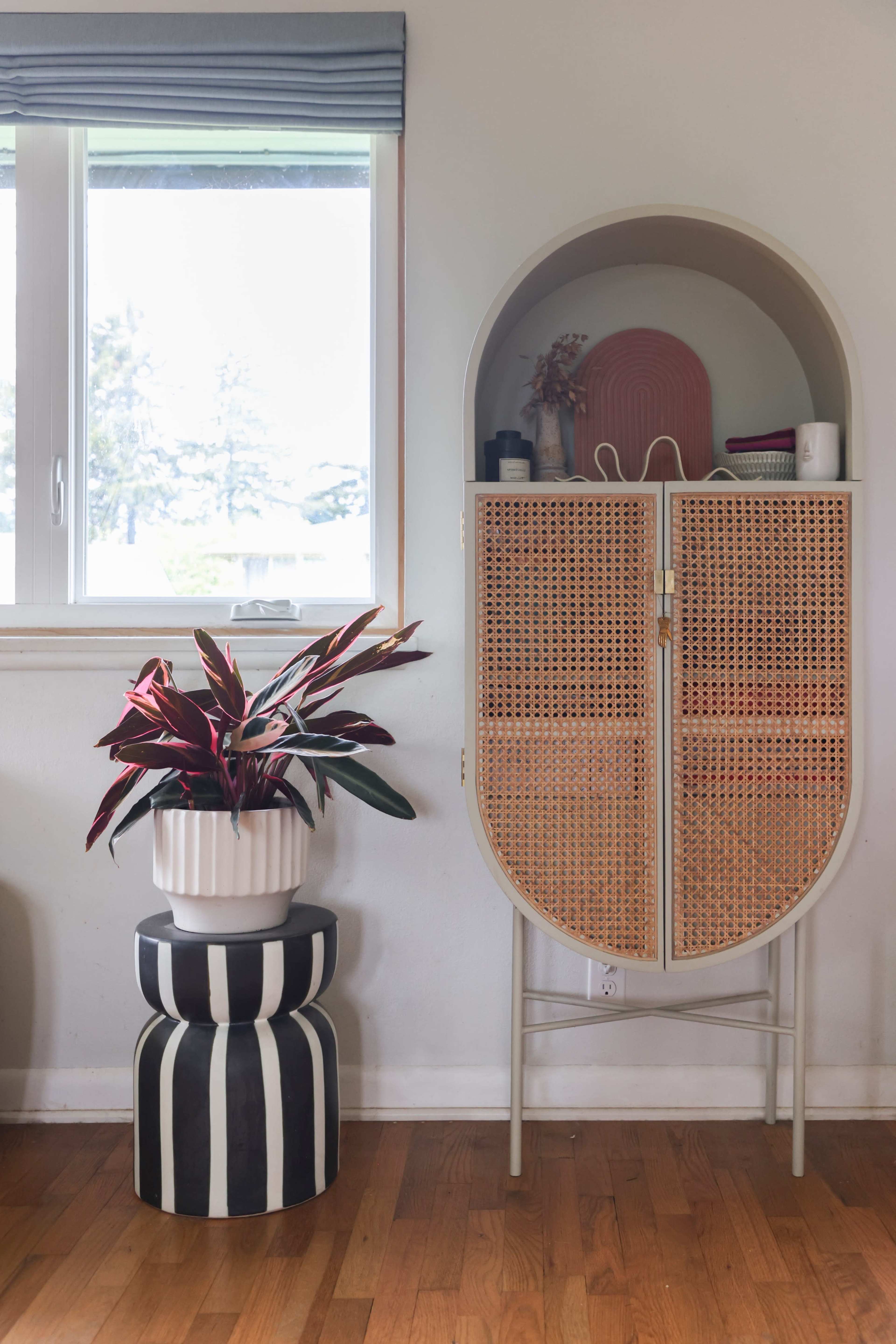 A rattan cabinet with closed doors stands next to a window, beside a striped stool topped with a white planter containing a red-leafed plant.