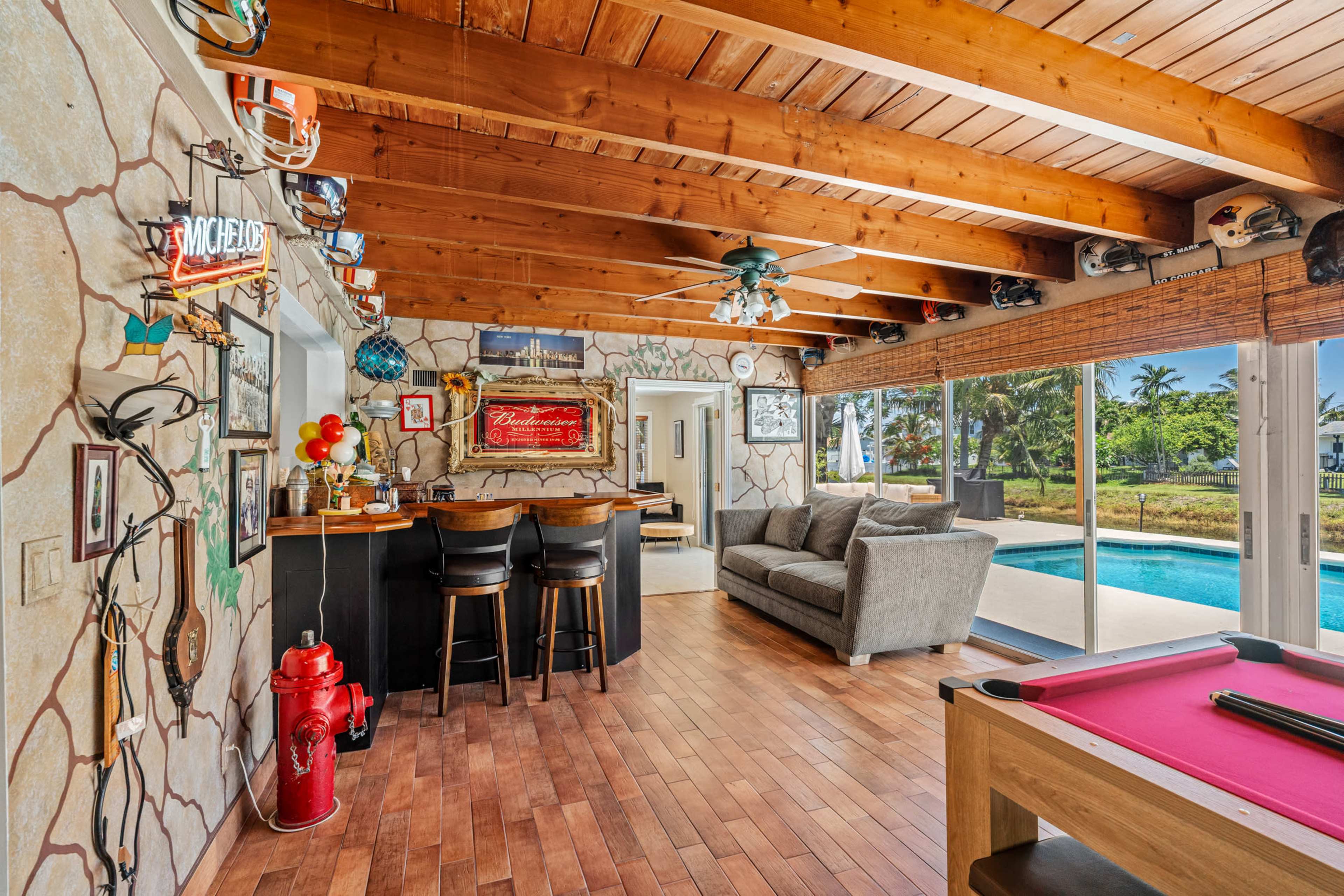 The image shows a spacious interior with a bar area, pool table, and large windows overlooking a pool and outdoor scenery, all under wooden beams.