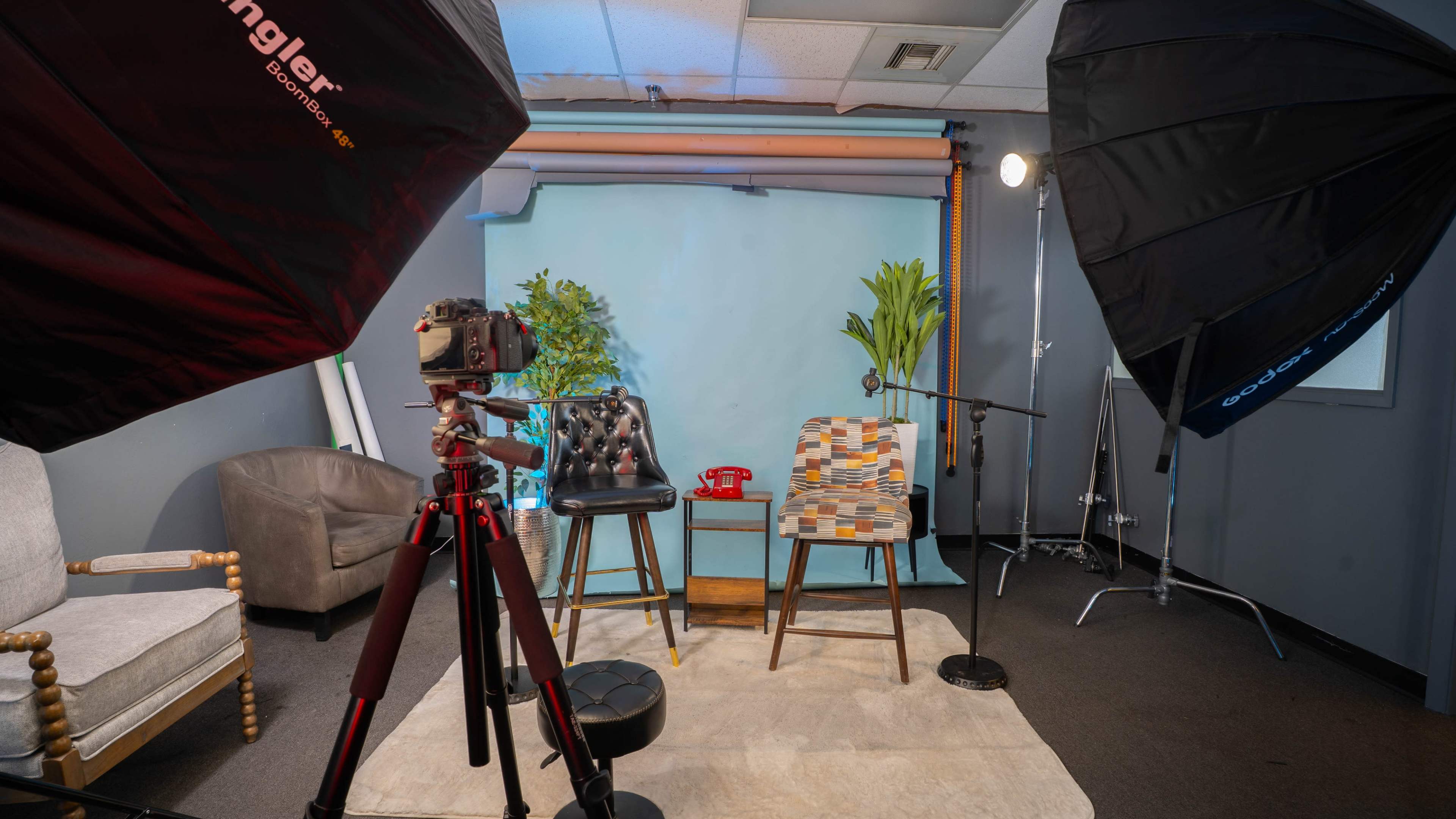 The image shows a photography setup featuring two chairs, a camera on a tripod, and softbox lights in a studio with a blue backdrop.
