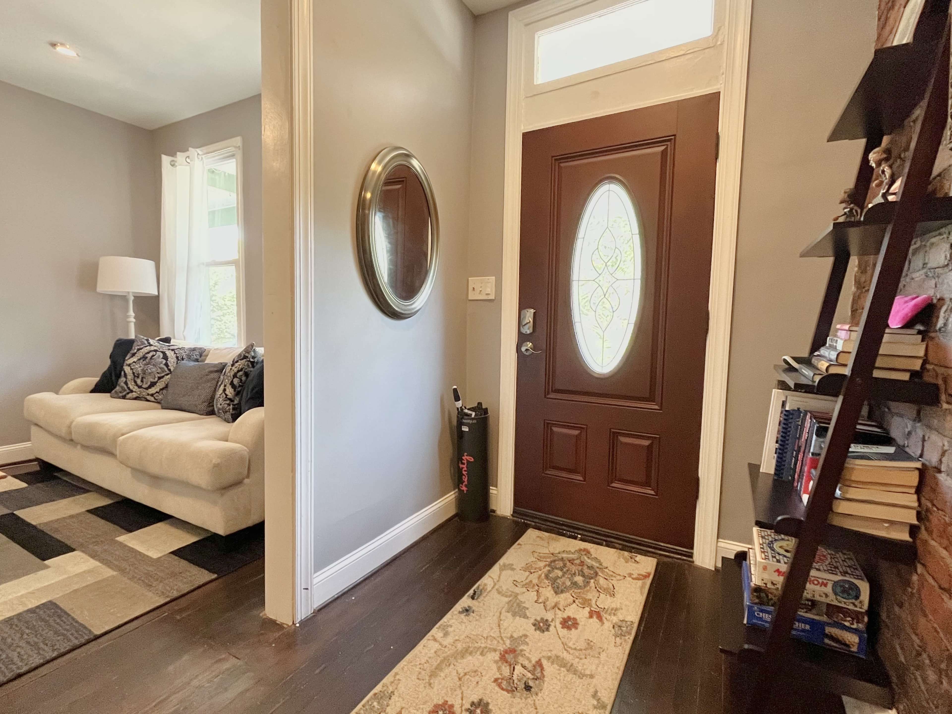 An entryway featuring a brown door with a decorative glass panel, a circular mirror on the wall, a beige sofa with decorative pillows, and a bookshelf filled with books.