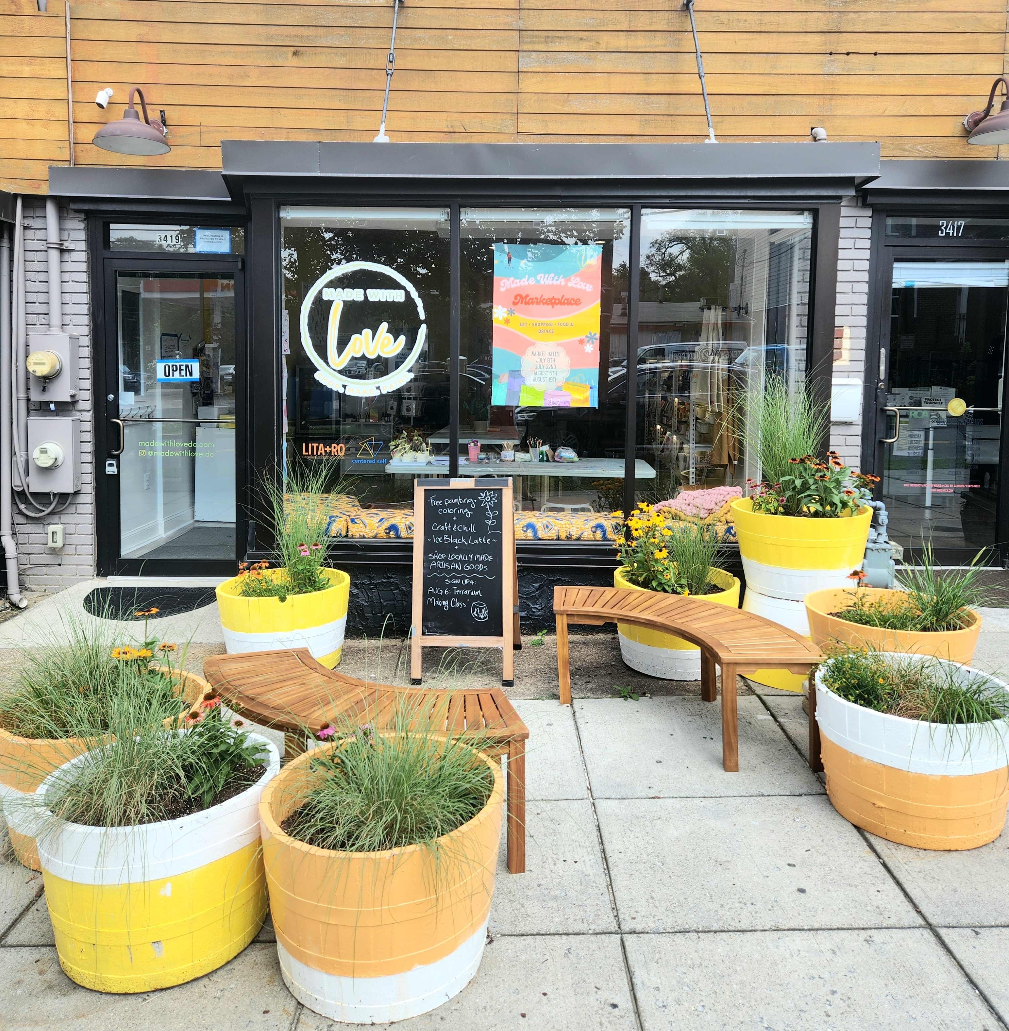 A storefront features large windows with an "Open" sign and planters filled with greenery and flowers arranged around wooden seating.